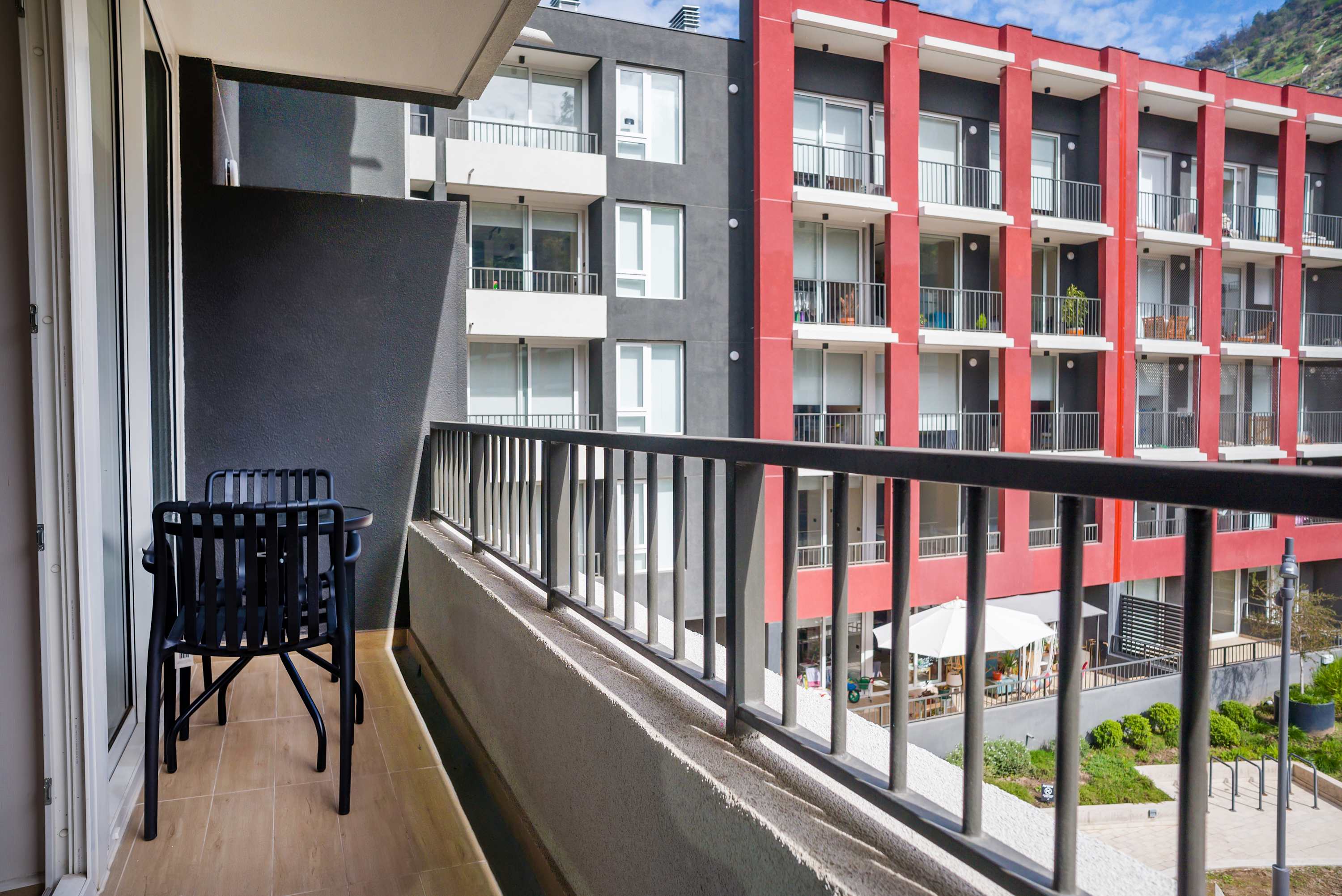 Private balcony with two chairs overlooking the landscaped courtyard, modern apartment buildings, and a green hillside beyond.
