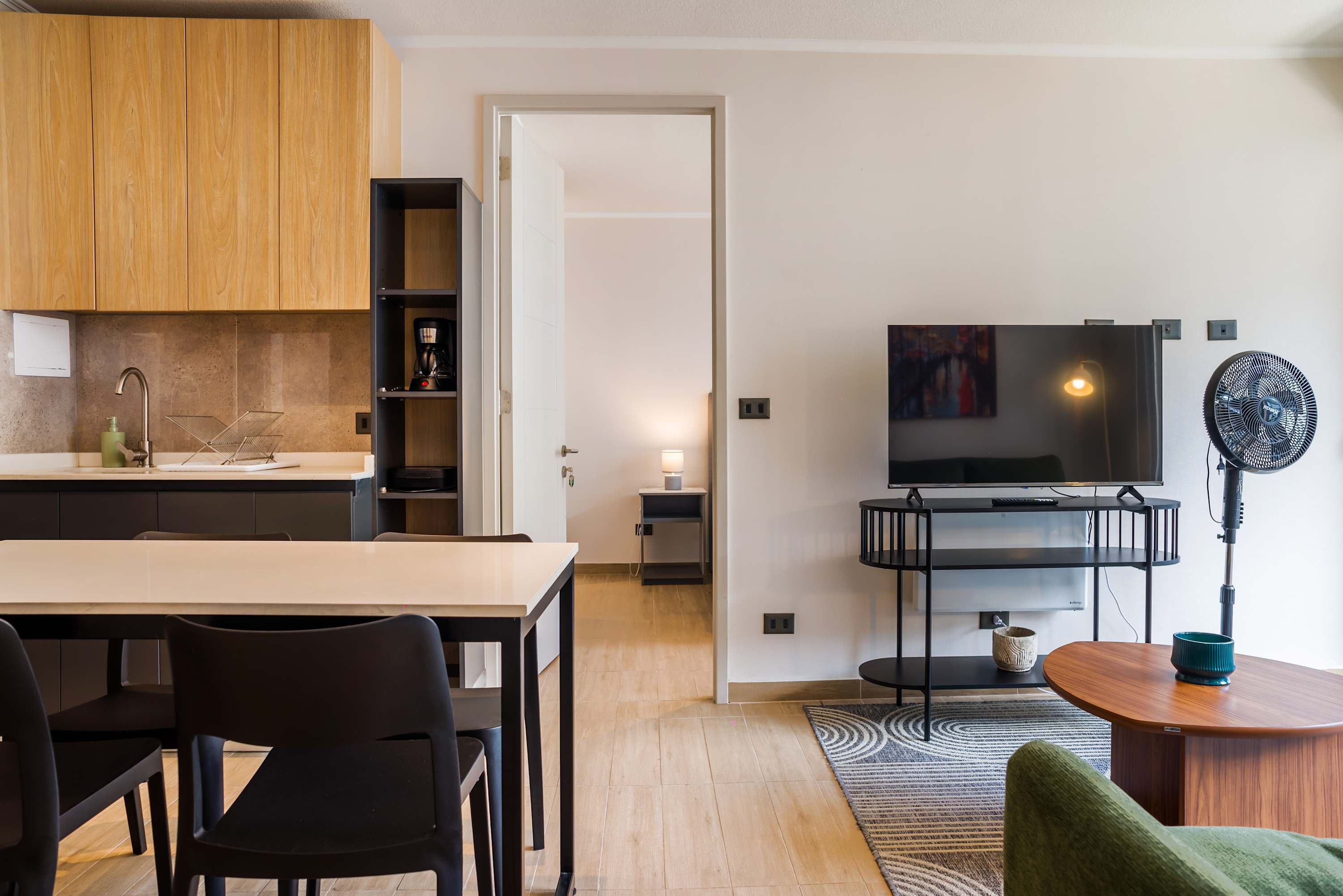 Open-plan living area with a fully equipped wood-accented kitchen, dining seating, and a large TV beside a patterned rug.
