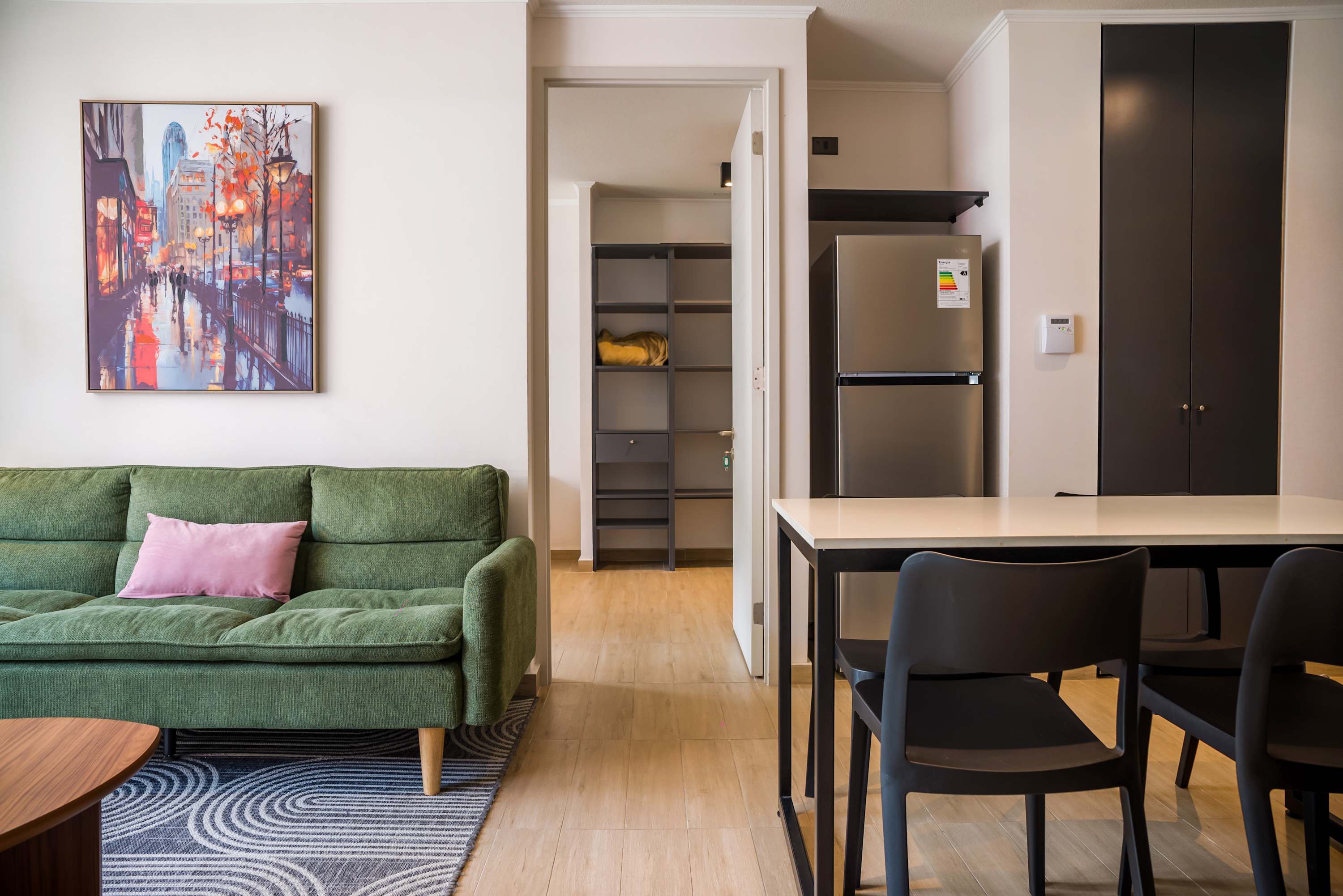 Open-plan living and dining area with a comfortable sage-green sofa, colorful cityscape artwork, and seating for shared meals.