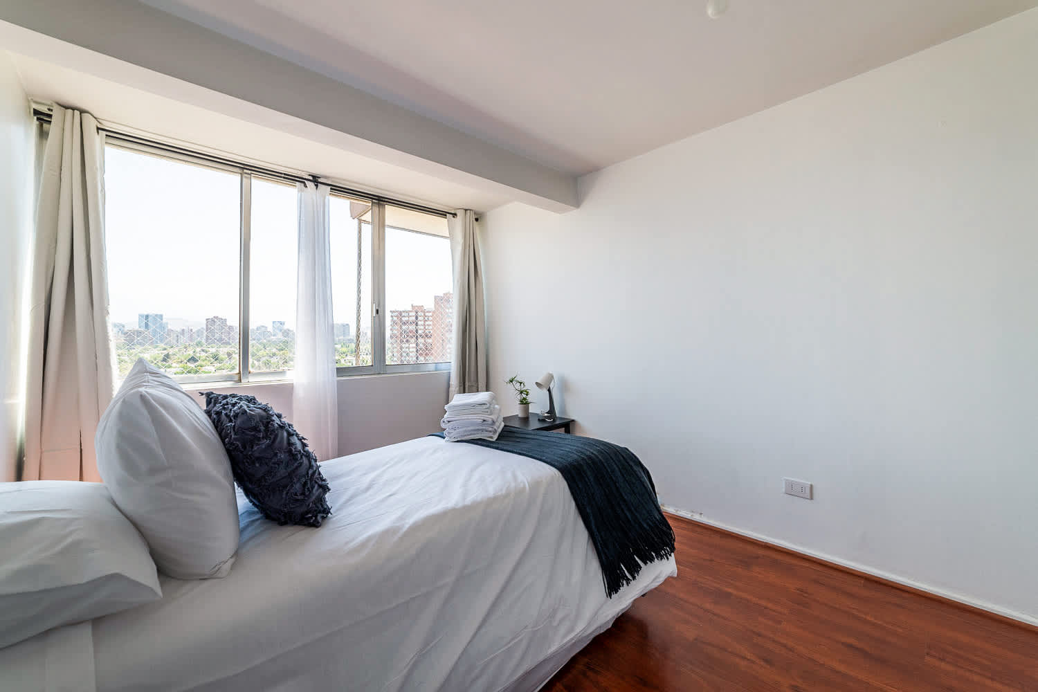 Bright bedroom with two neatly made beds, a cozy charcoal throw, and large windows overlooking the city and surrounding greenery.