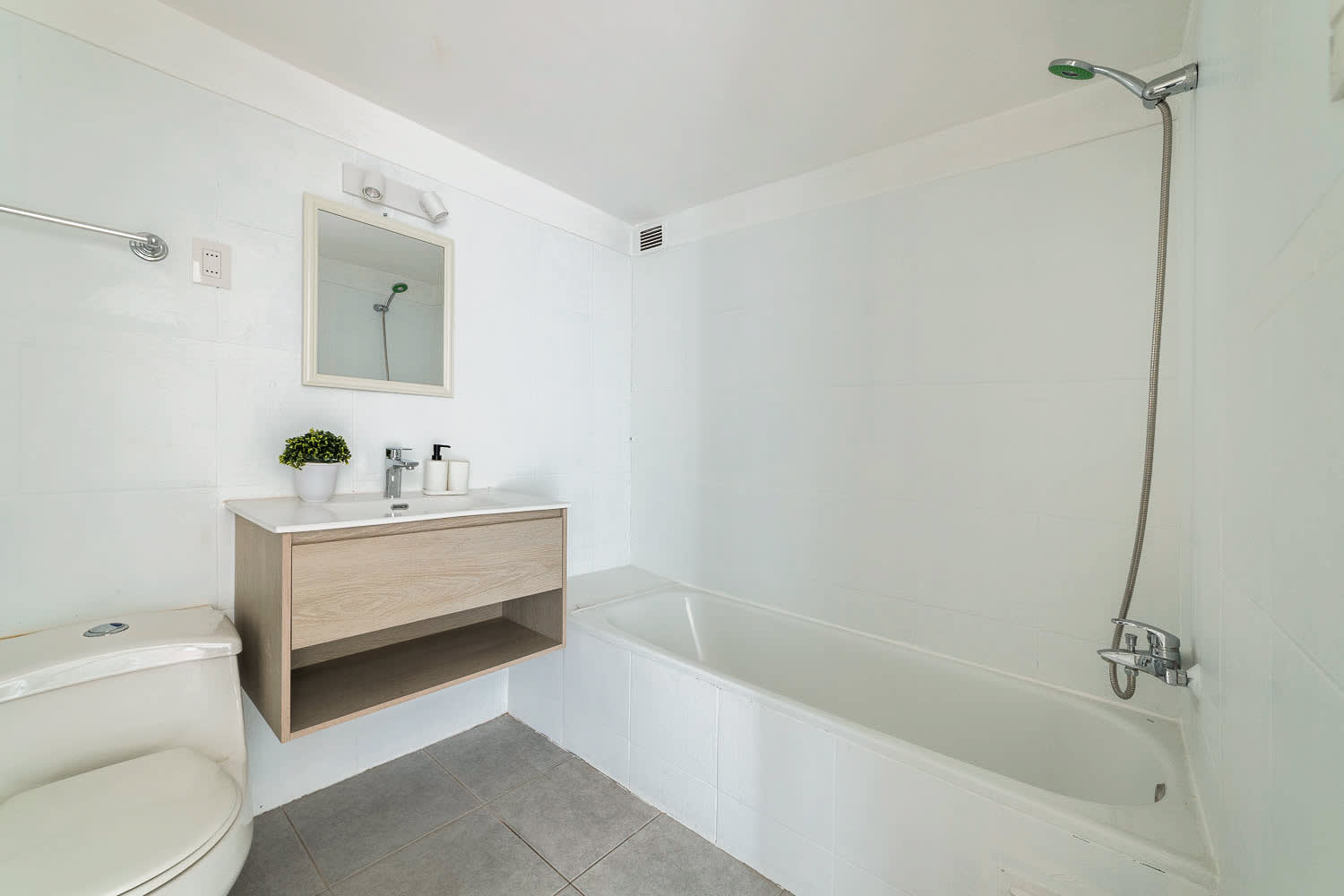 Bright private bathroom with a full-size bathtub, handheld shower, light wood vanity, framed mirror, and clean white tile finishes.