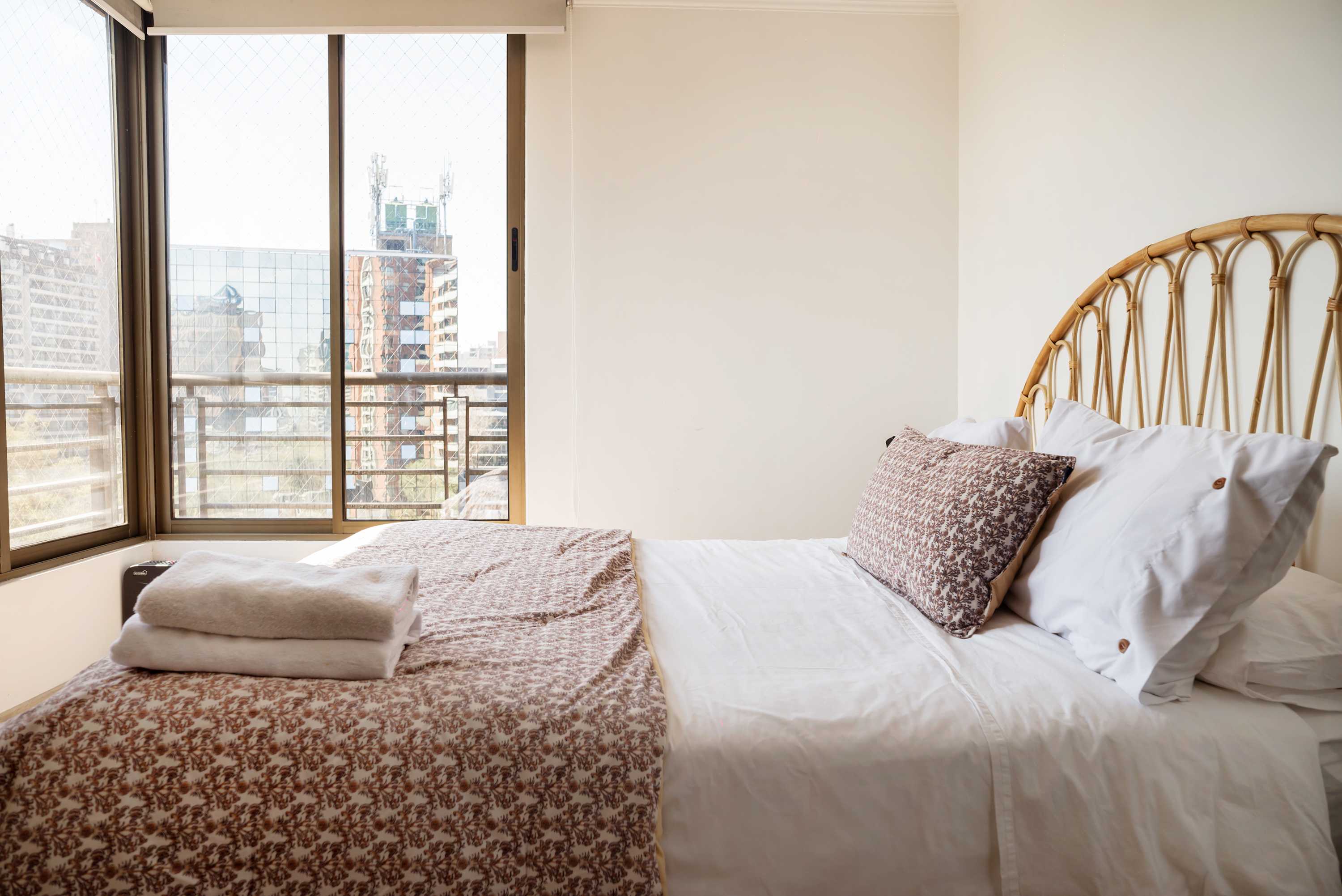 Bright bedroom with two neatly made beds, a natural rattan headboard, and expansive city views through the corner windows.