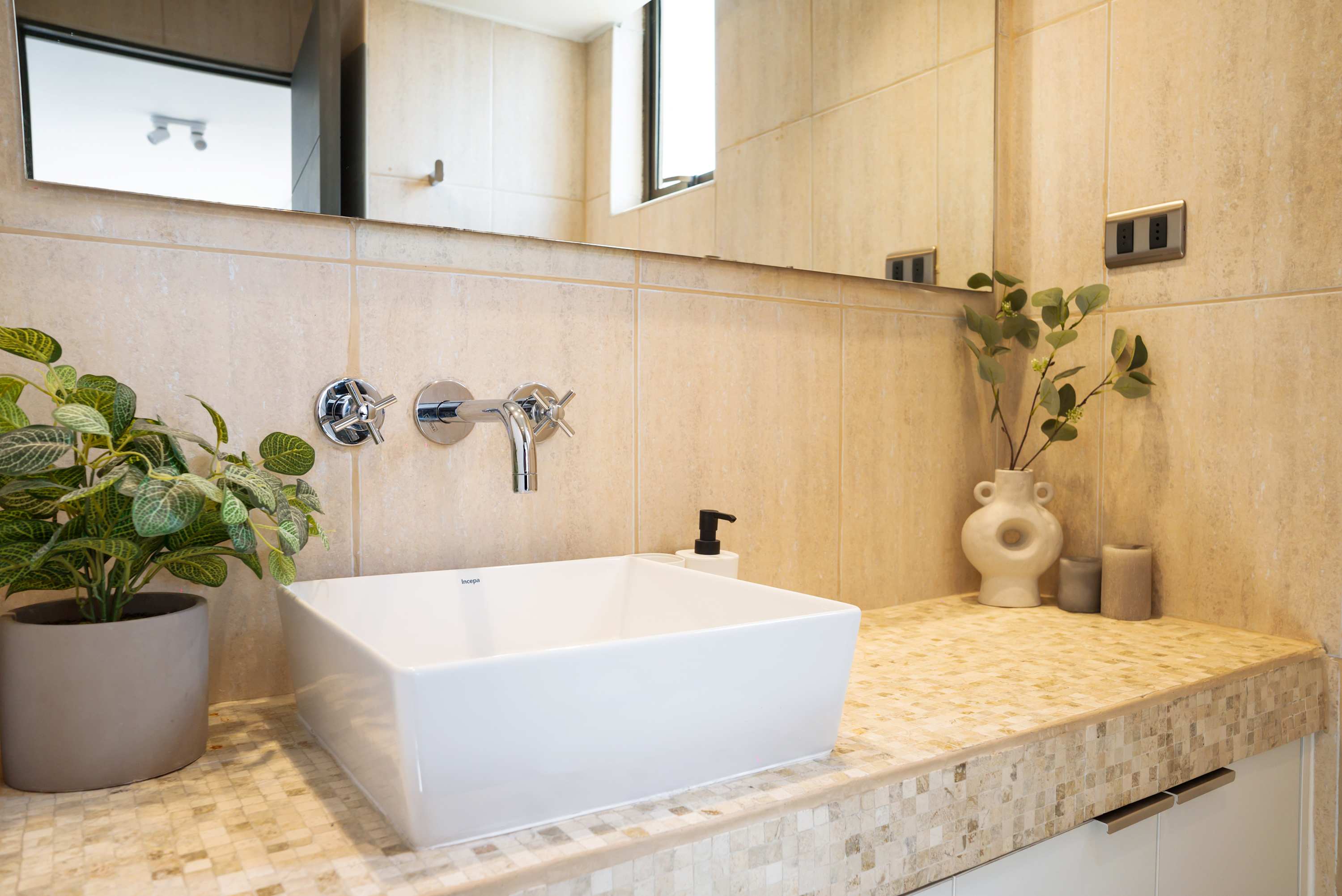 Contemporary bathroom with a sculptural white basin, wall-mounted chrome faucet, warm stone-look tiles, and greenery beside a bright mirror.