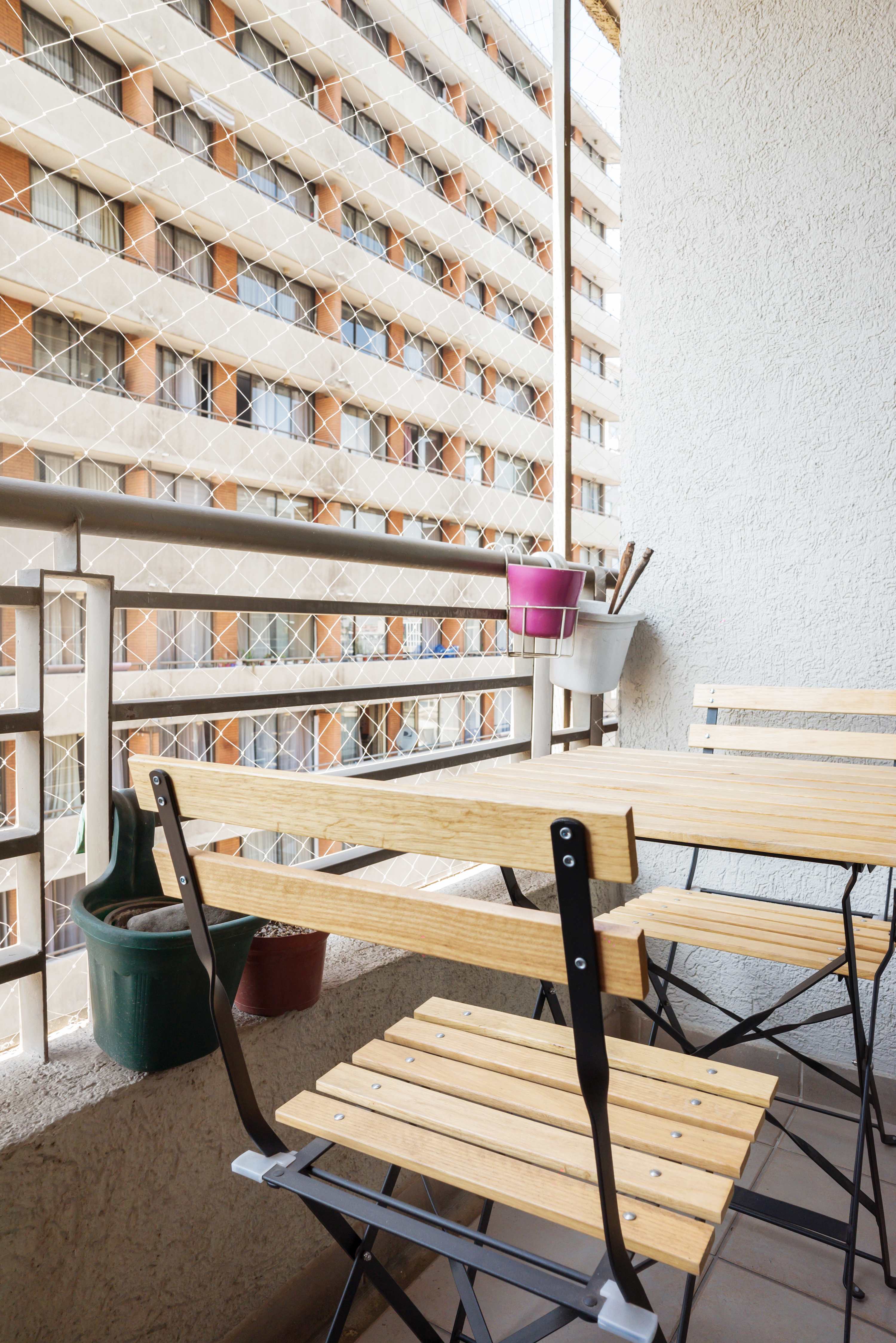 Private balcony with a wooden dining set, potted plants, and protective safety netting overlooking the surrounding apartment buildings.