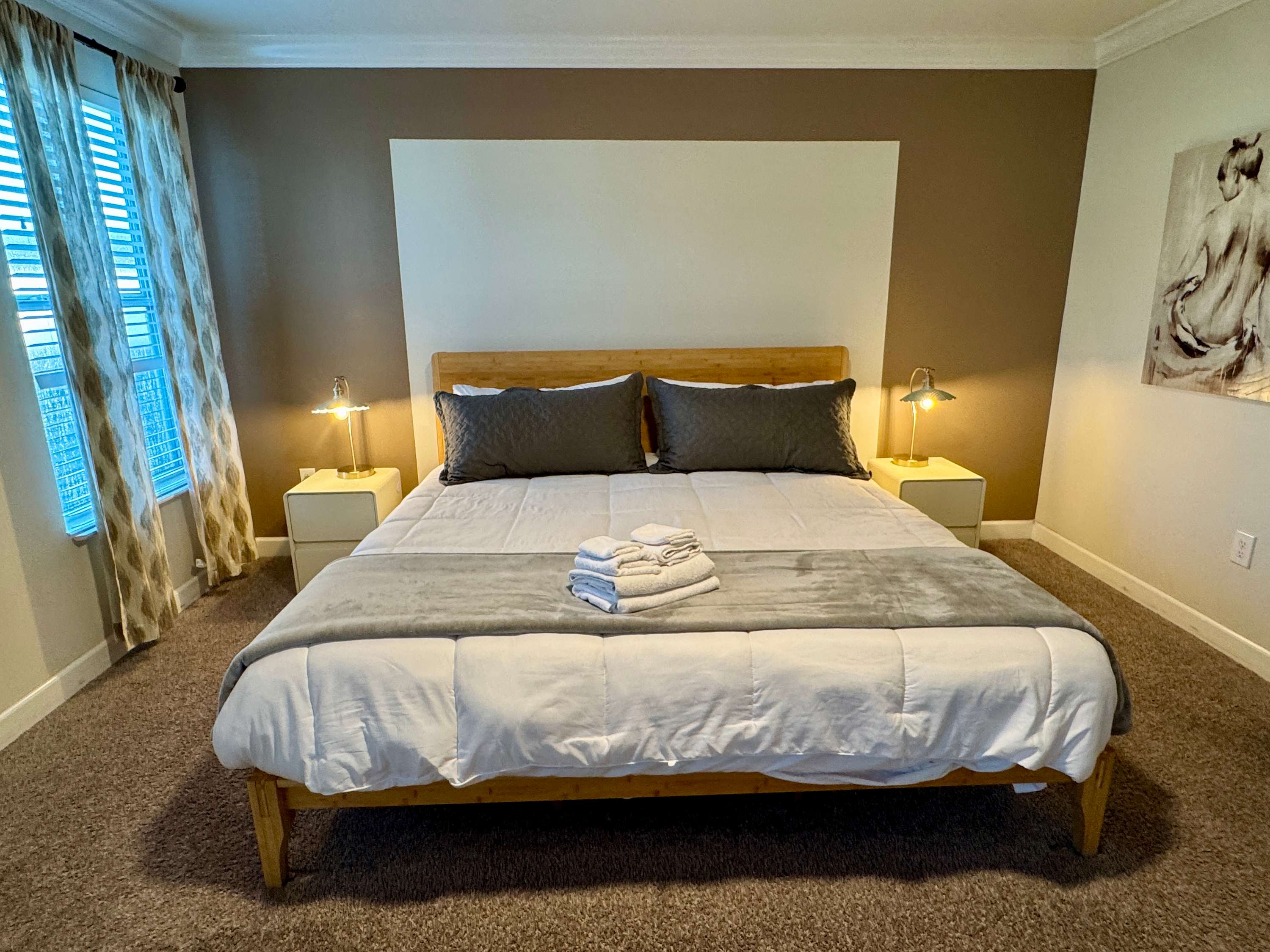 Comfortable bedroom with a light wood headboard, soft gray bedding, matching bedside lamps, and a bright window with patterned curtains.