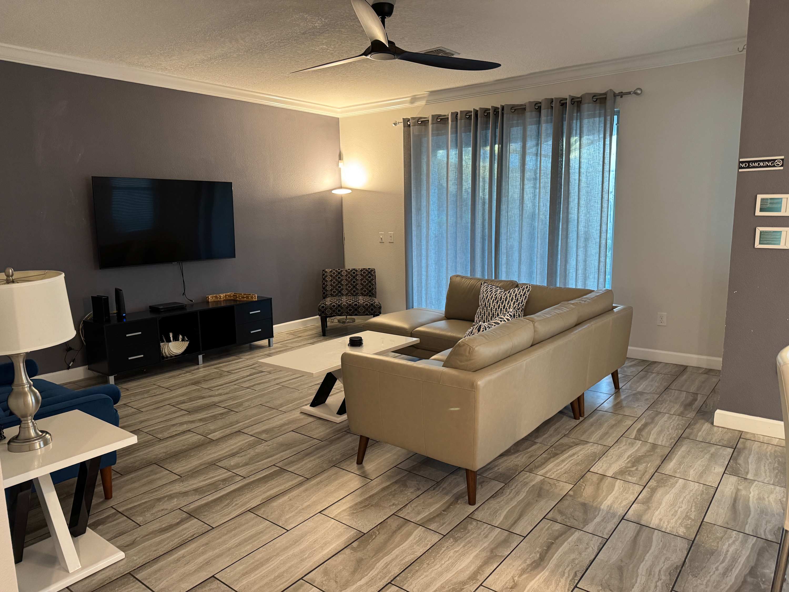 Spacious living room with a large leather sectional, flat-screen TV, and filtered daylight through floor-to-ceiling curtains.