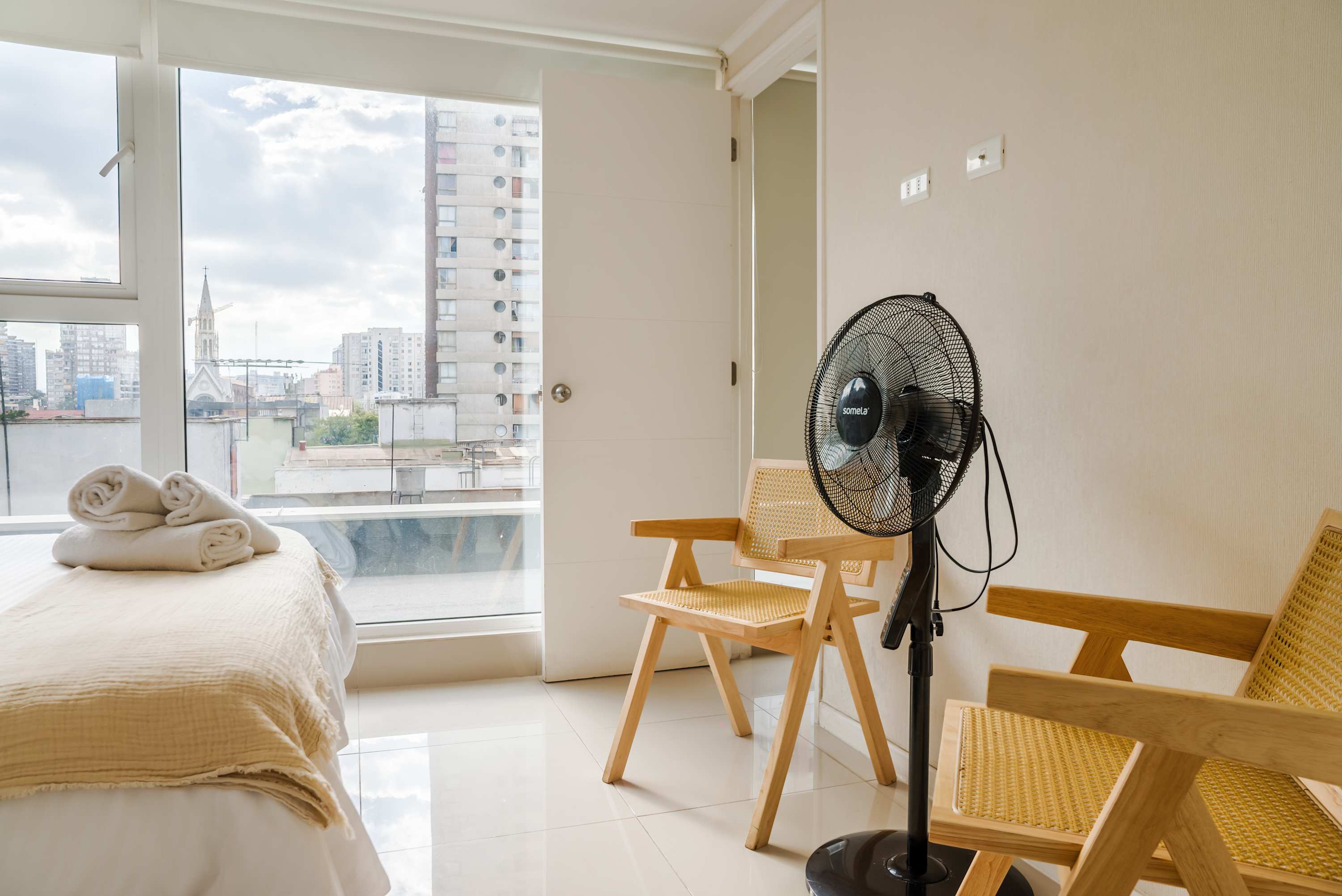 Bright studio bedroom with a neatly made bed, folded towels, and expansive city views featuring a distinctive church steeple.