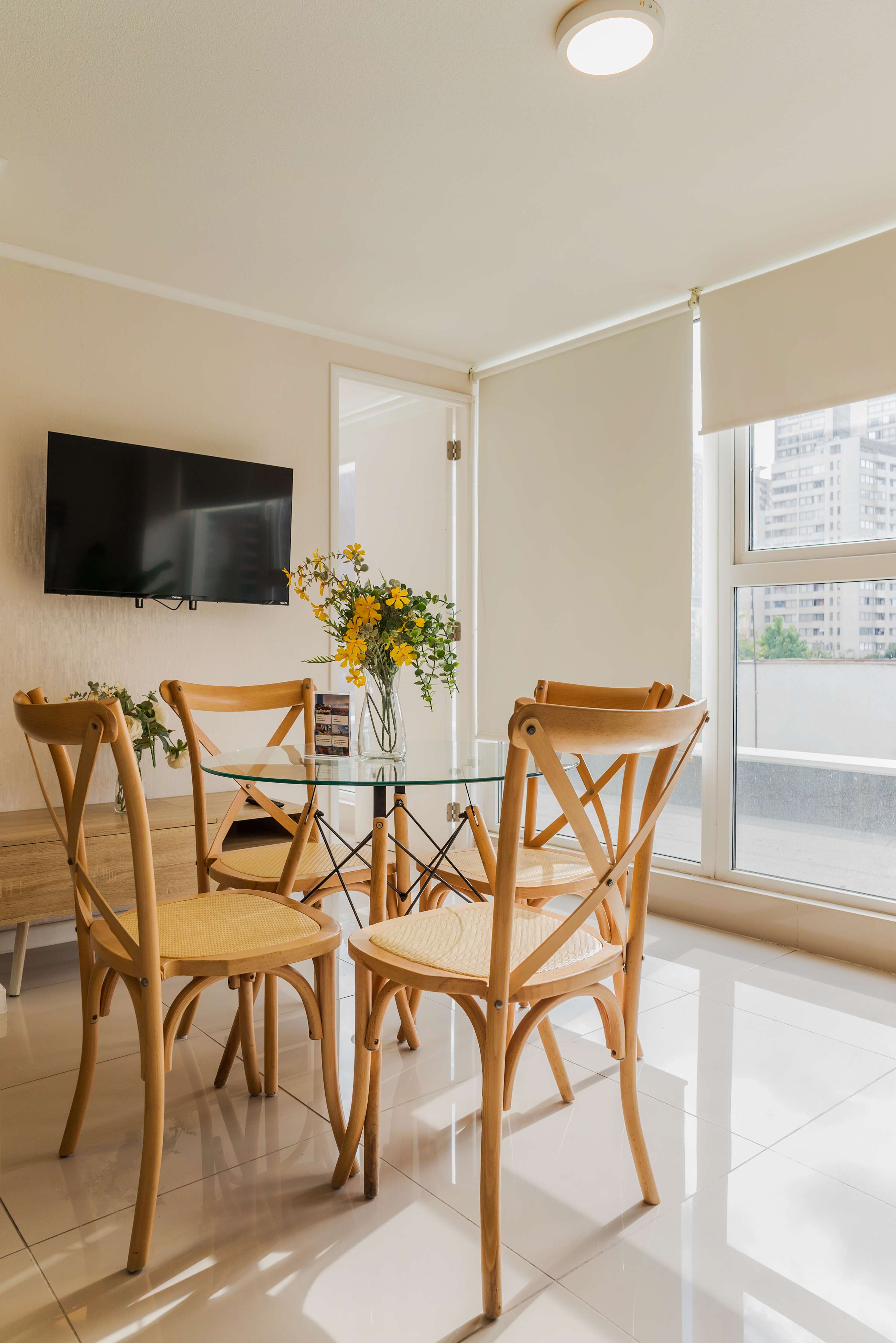 Private dining area with a round glass table, warm wood chairs, yellow flowers, and bright city views through the floor-to-ceiling windows.