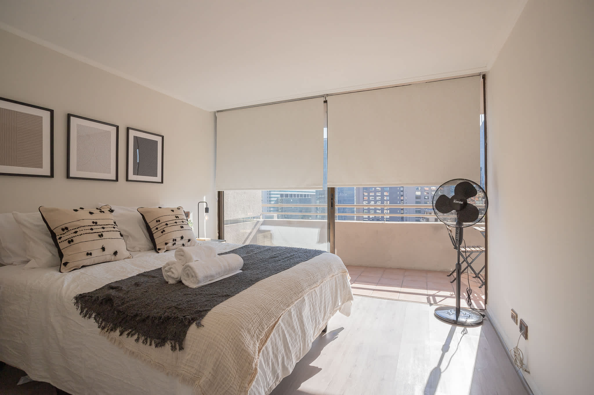 Bright bedroom with crisp white bedding, folded towels, and large windows opening to a private balcony with city views.