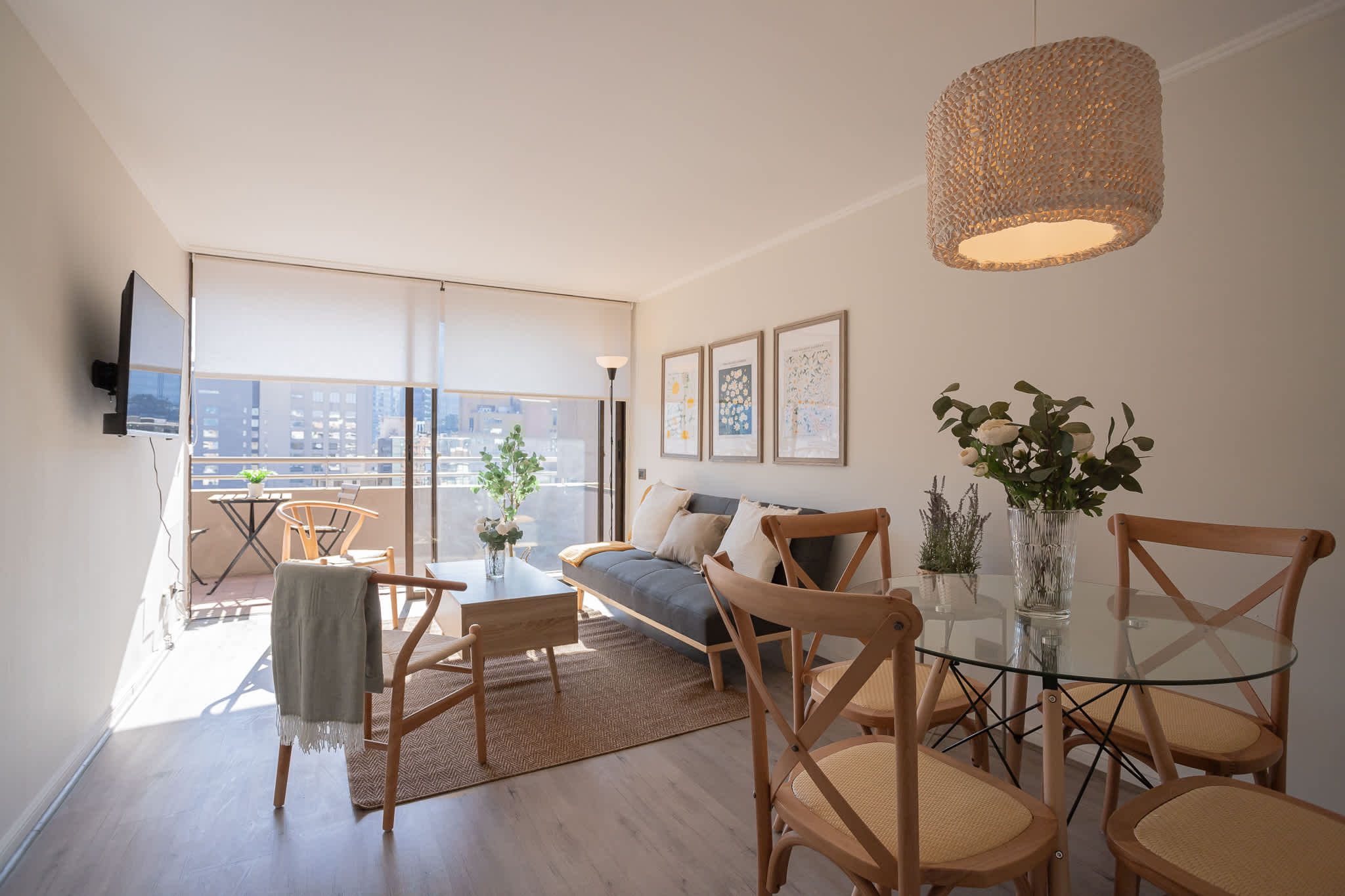 Sunlit living and dining area with a comfortable sofa, woven pendant light, and balcony overlooking the city.