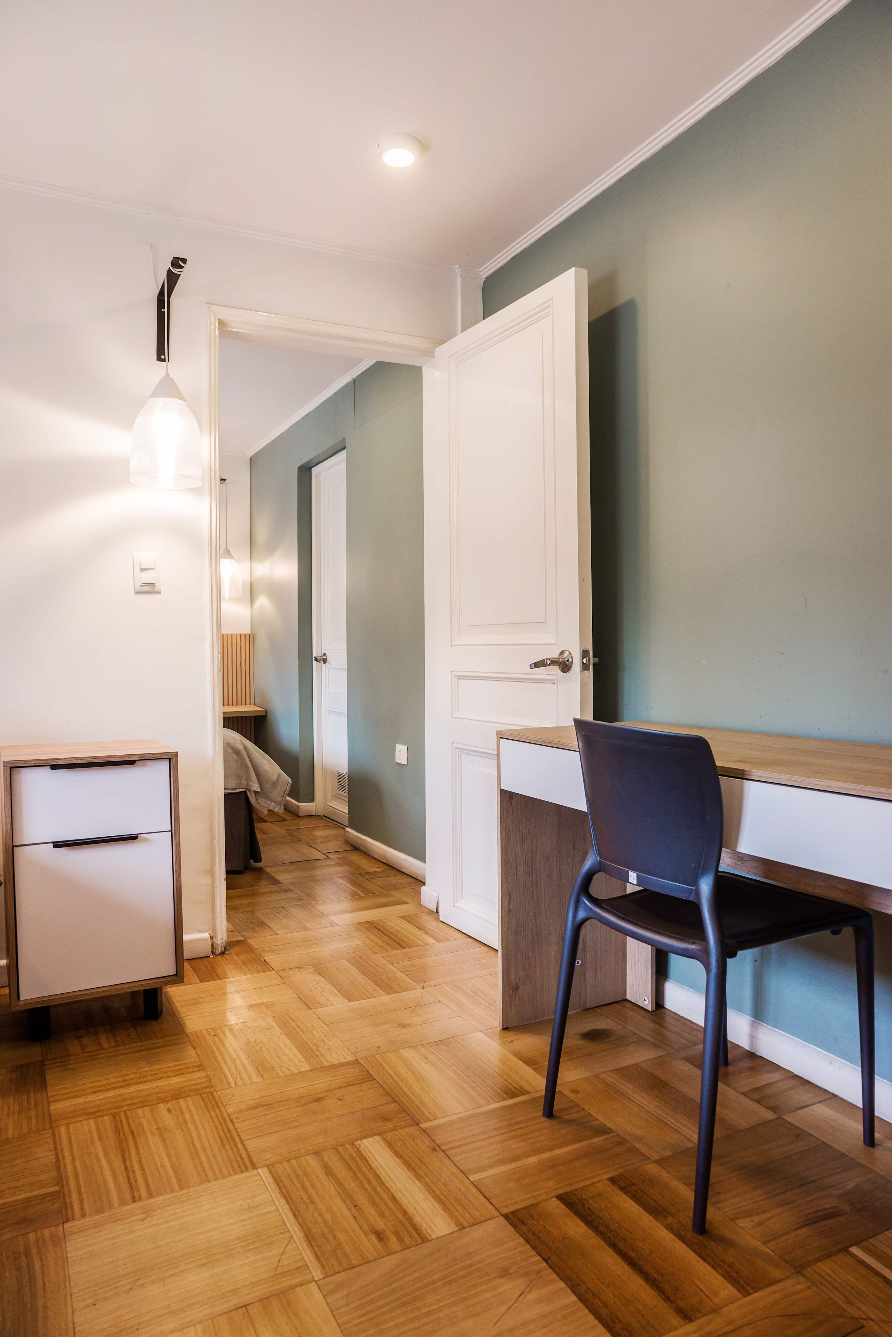 Compact apartment workspace with a wood desk, comfortable chair, and warm parquet flooring beside the sleeping area.
