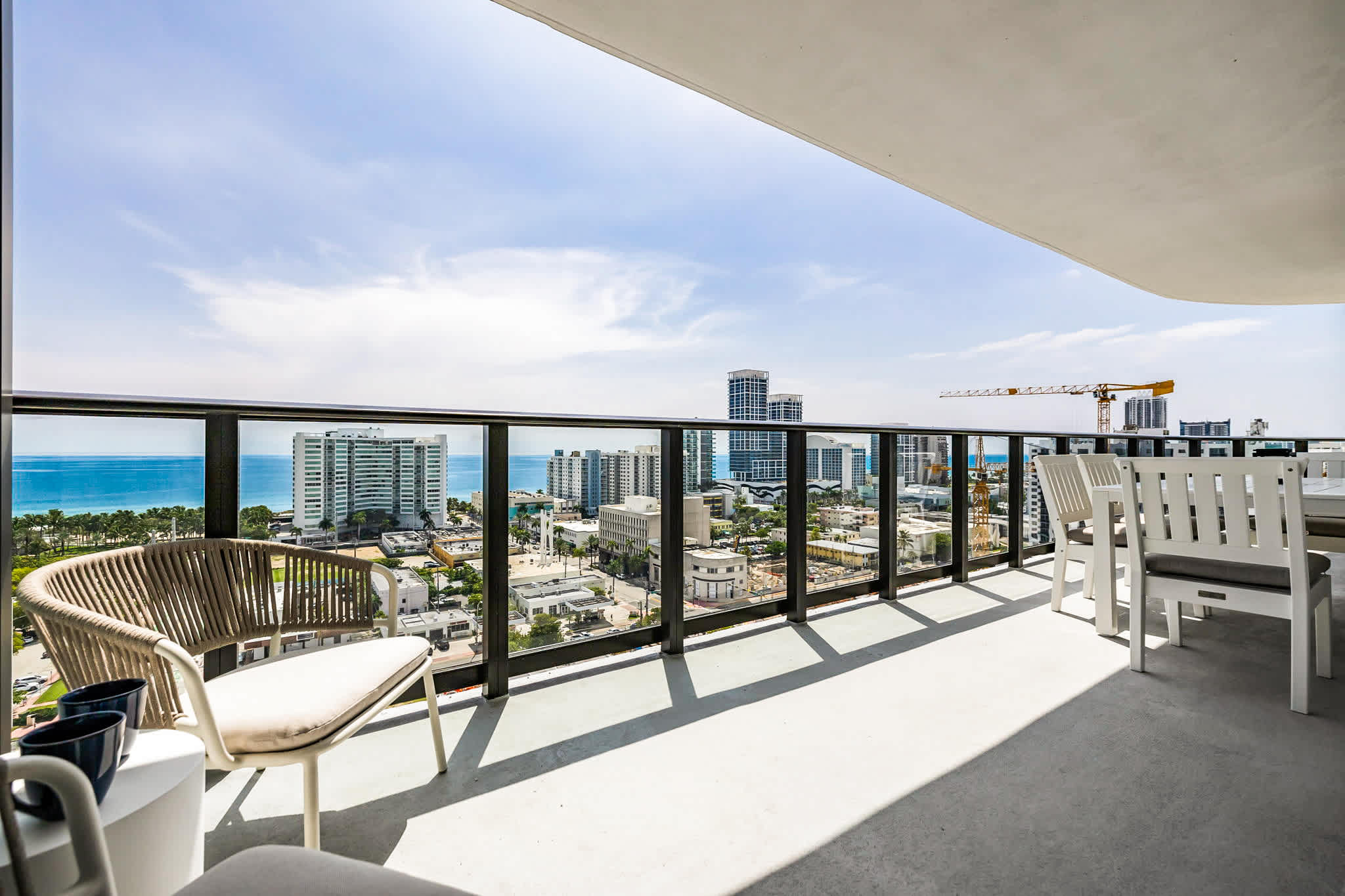 Private balcony with outdoor dining and lounge seating overlooking the blue water, palm-lined shore, and vibrant city skyline.