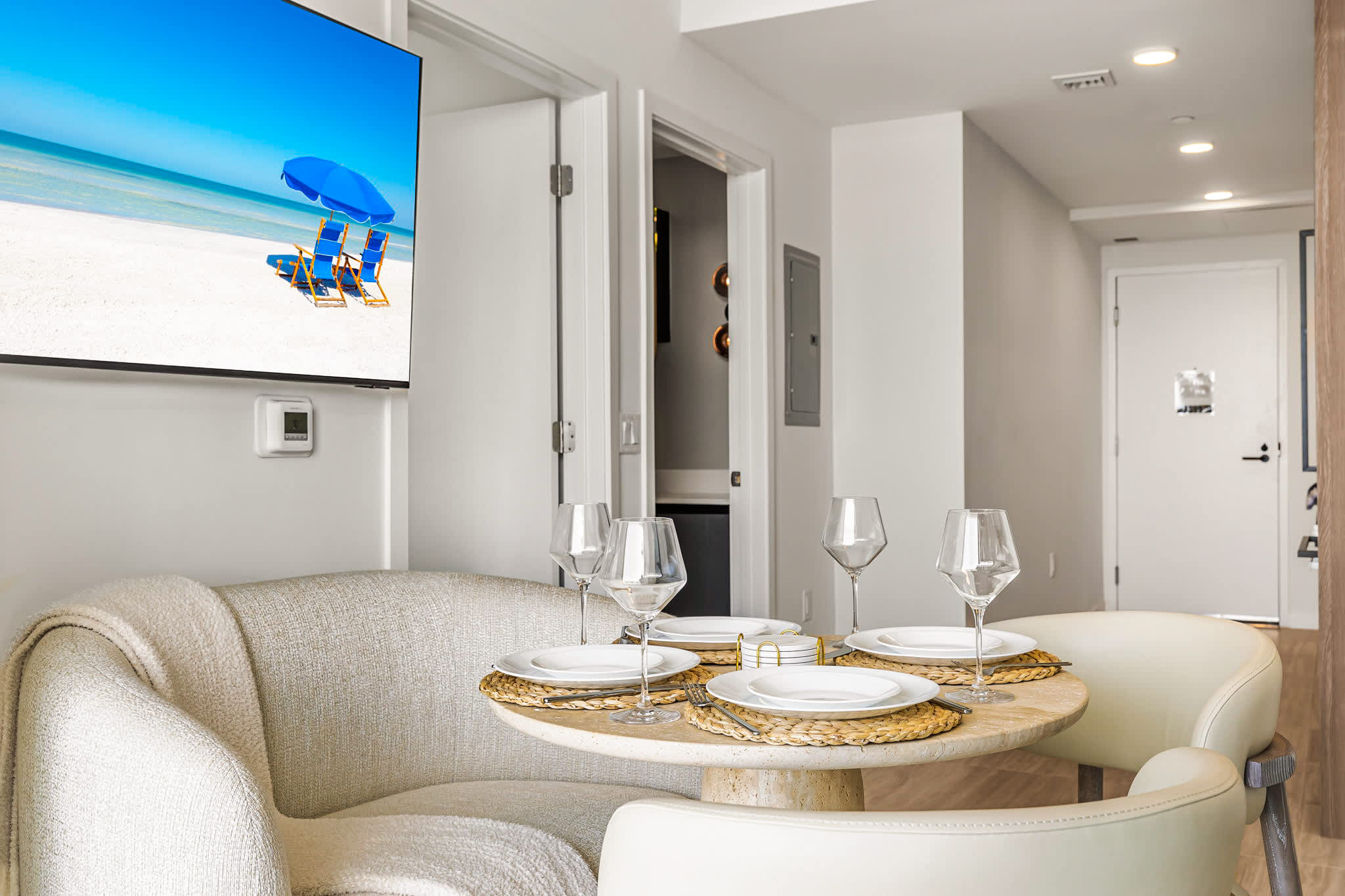 Private dining area with a round table set for four, woven placemats, wine glasses, and a large beach scene on the wall-mounted TV.