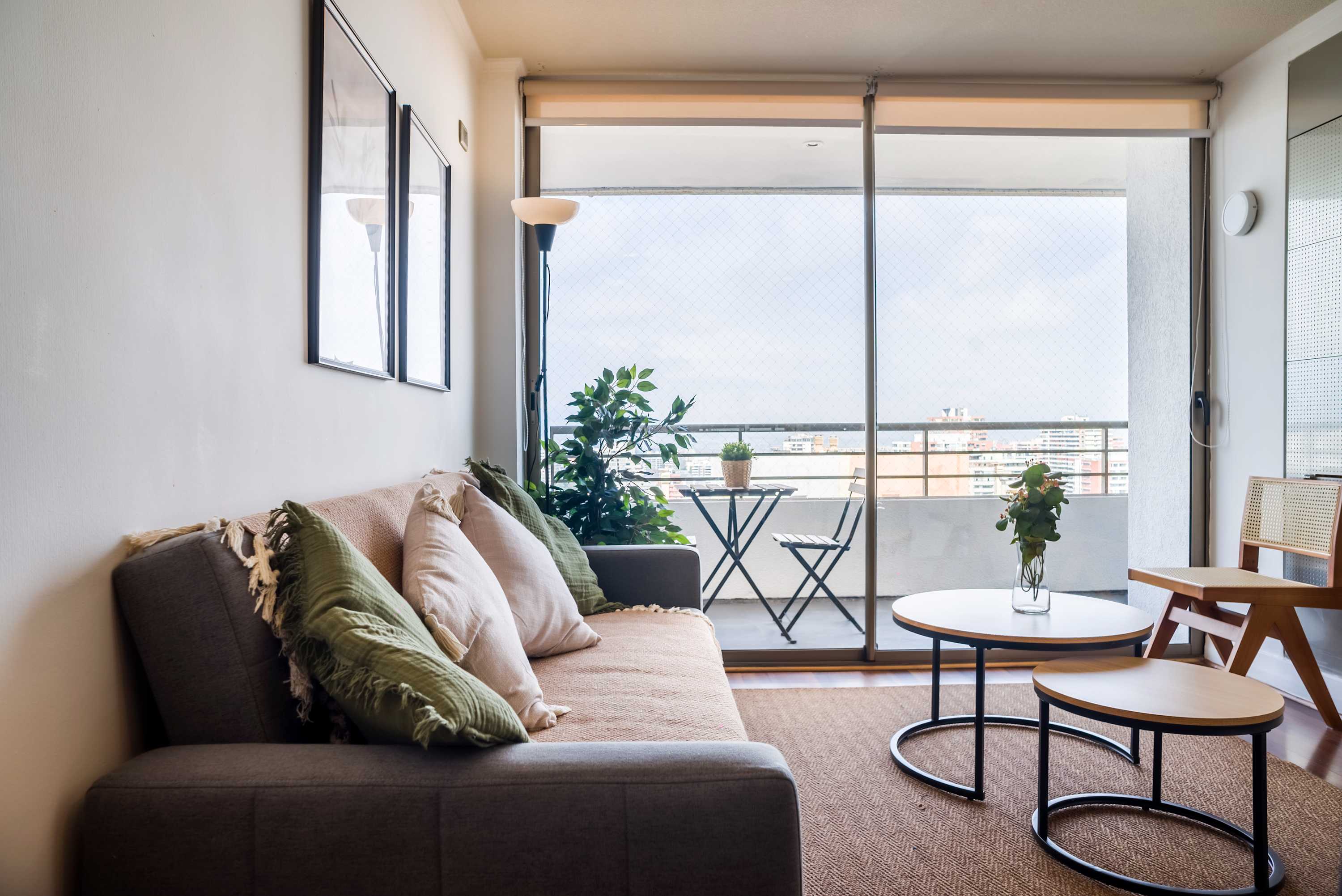 Bright living room with layered neutral textiles, leafy plants, and a furnished balcony overlooking the city.