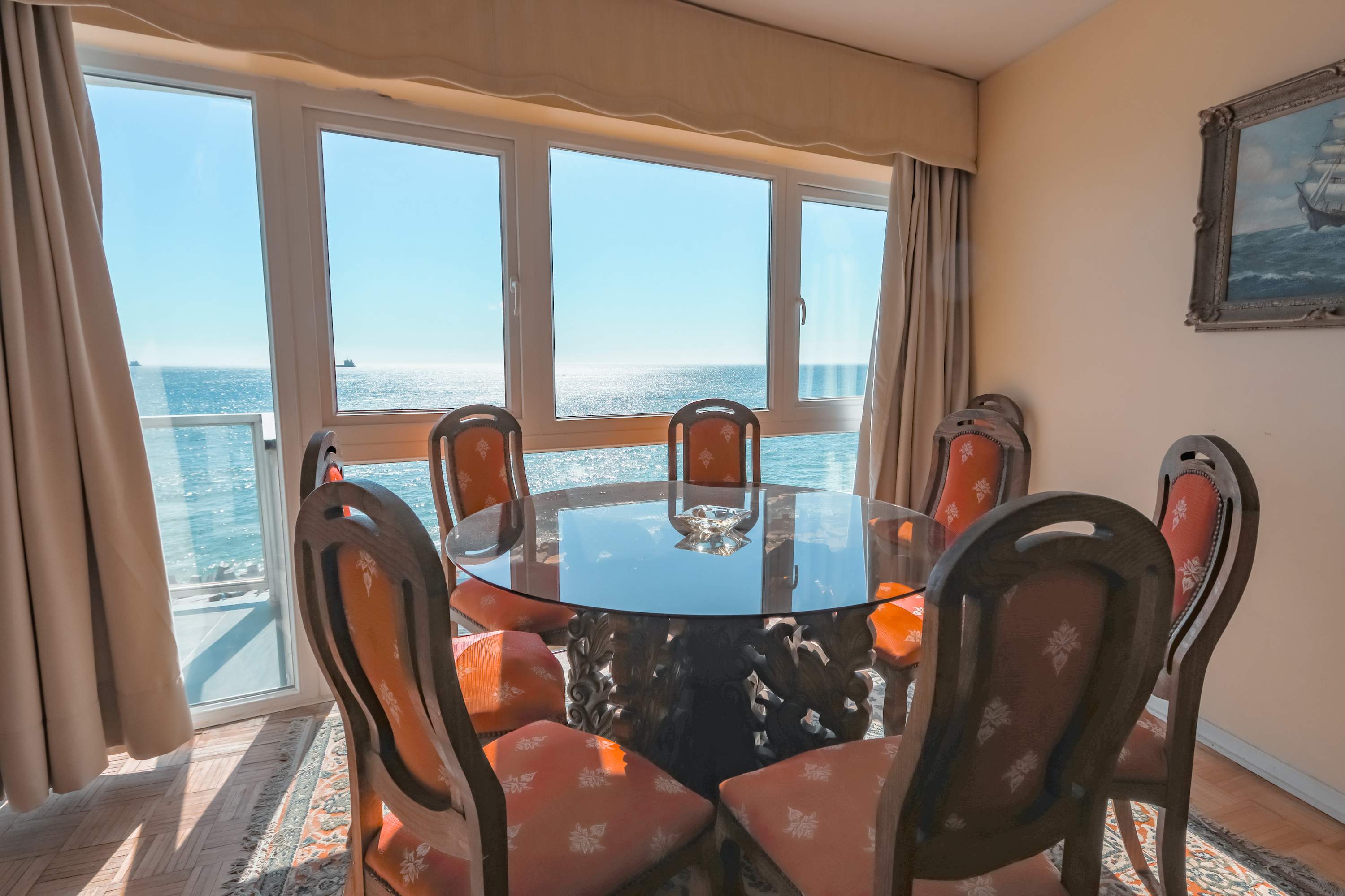 Elegant dining room with a round glass table, ornate seating, and expansive ocean views through floor-to-ceiling windows.