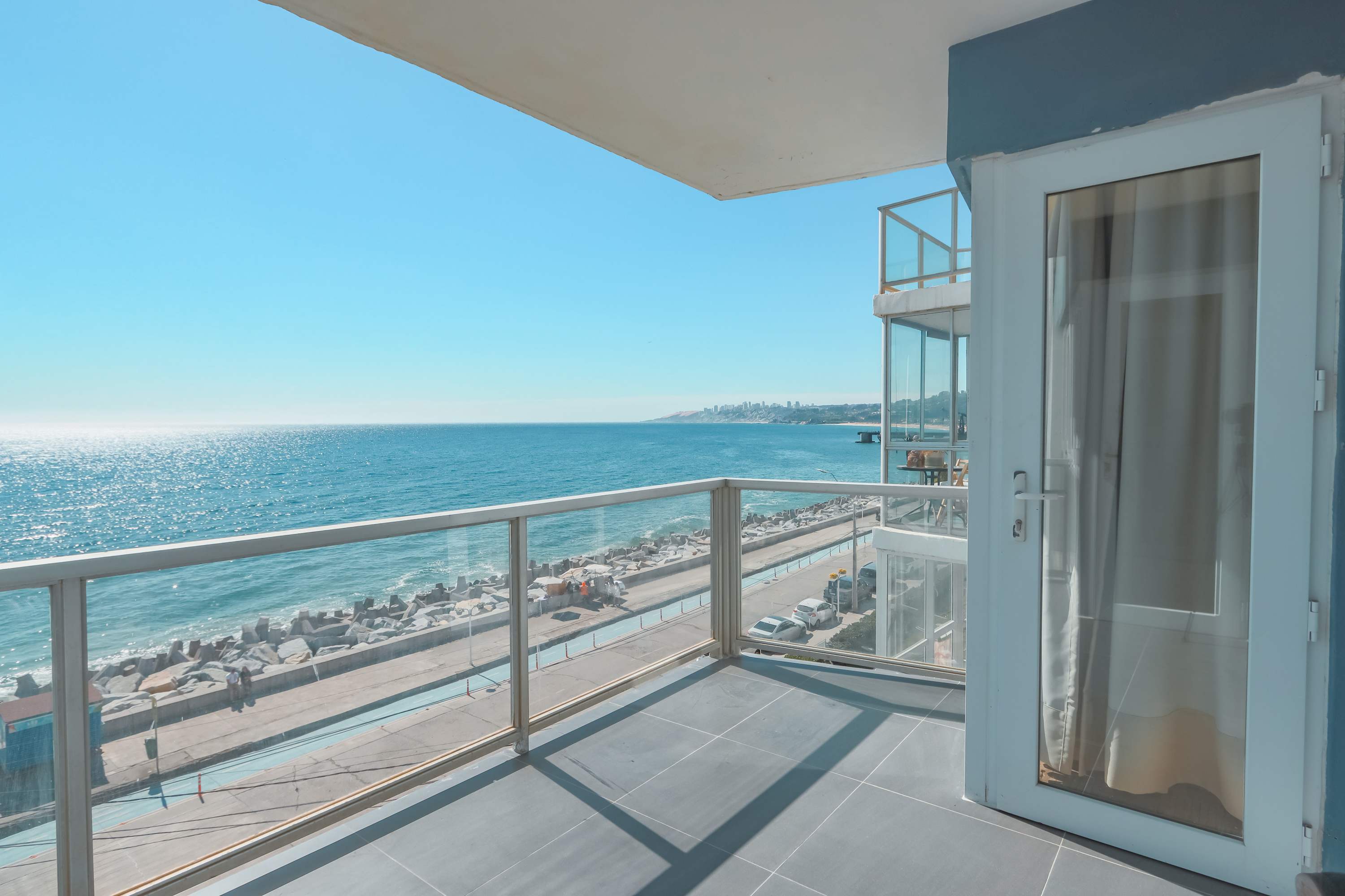 Private oceanfront balcony with floor-to-ceiling glass railings and sweeping views of the Pacific, rocky shoreline, and coastal promenade.