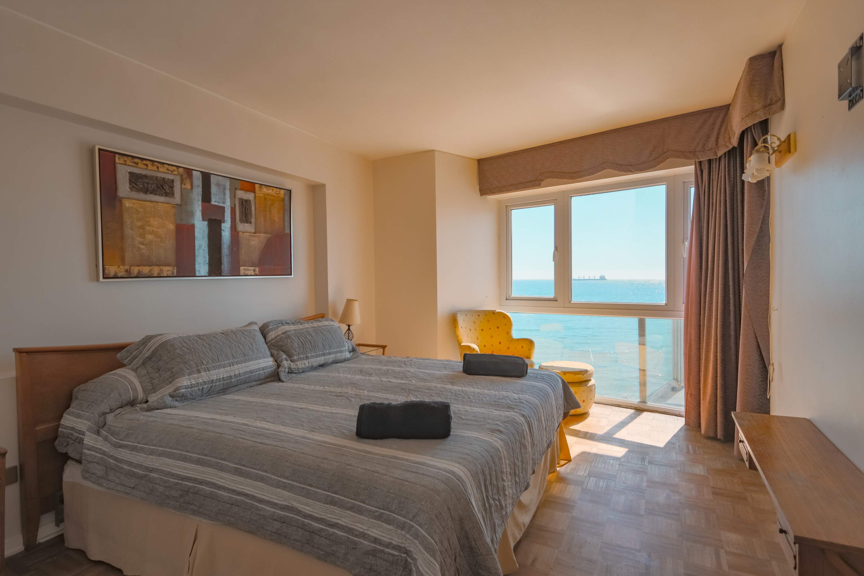 Ocean-view bedroom with a comfortable gray bed, colorful artwork, and a mustard armchair beside wide windows overlooking the blue water.