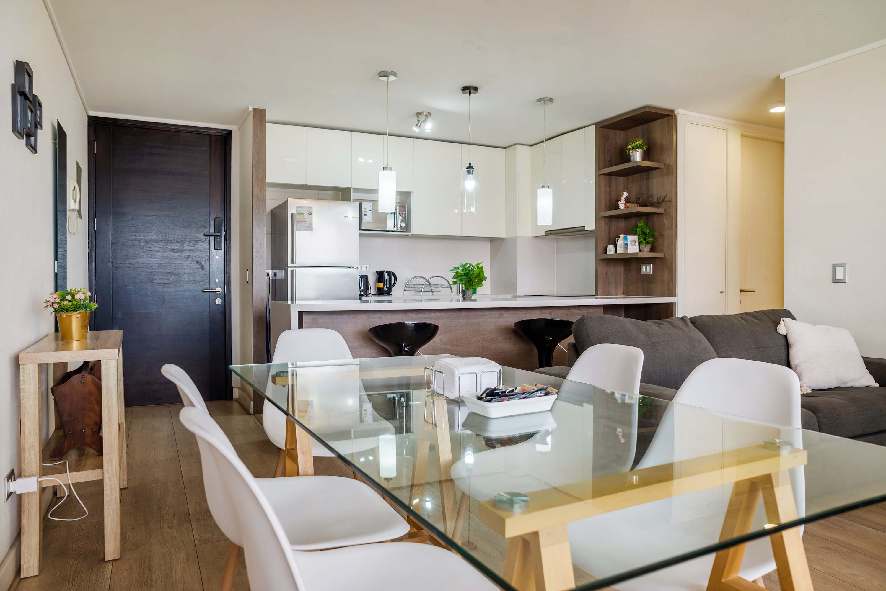 Open-plan living and dining area with a glass table, white chairs, gray sofa, and a modern fully equipped kitchen.