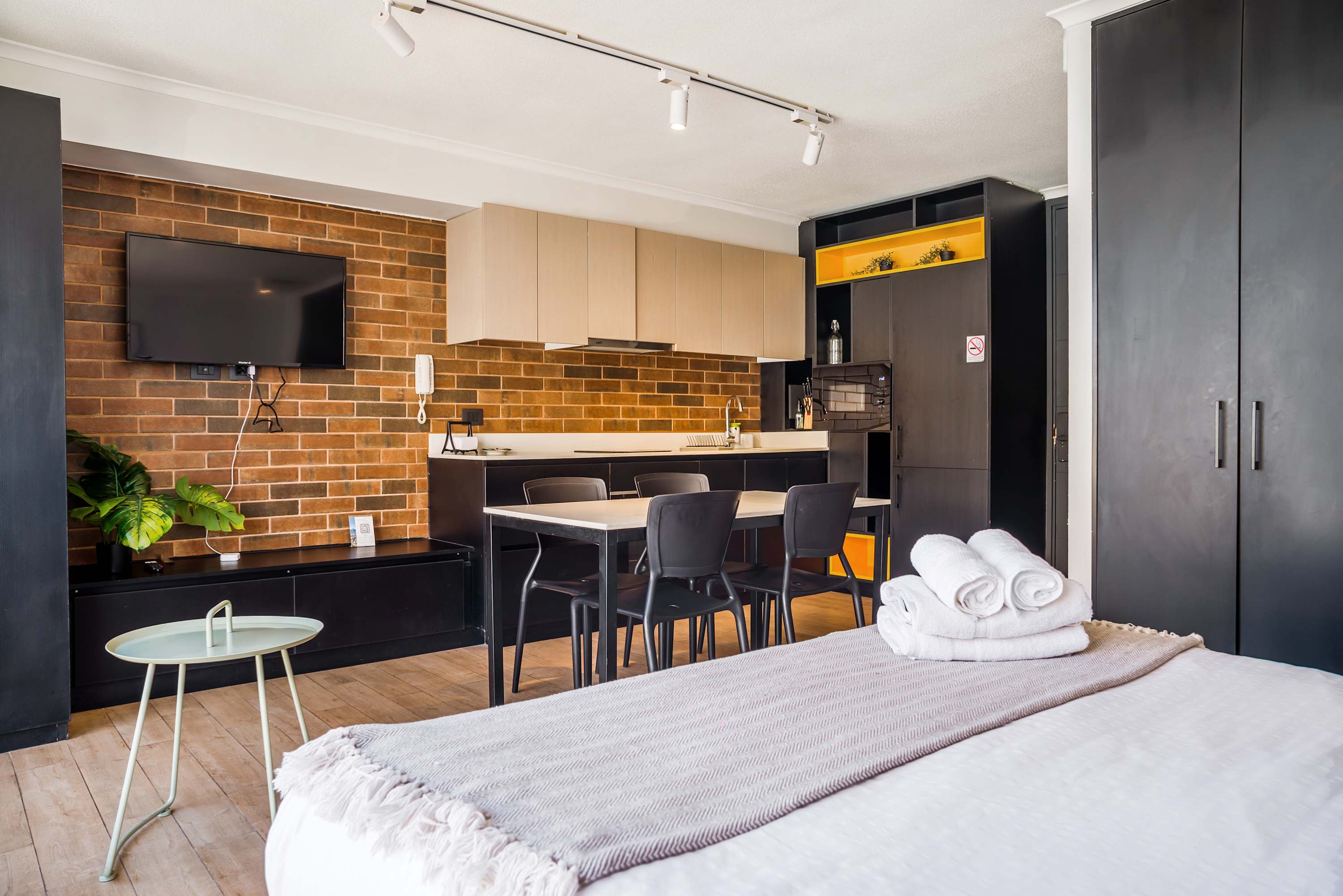 Private studio bedroom with a neatly made bed, exposed brick accent wall, compact kitchenette, and warm illuminated shelving.