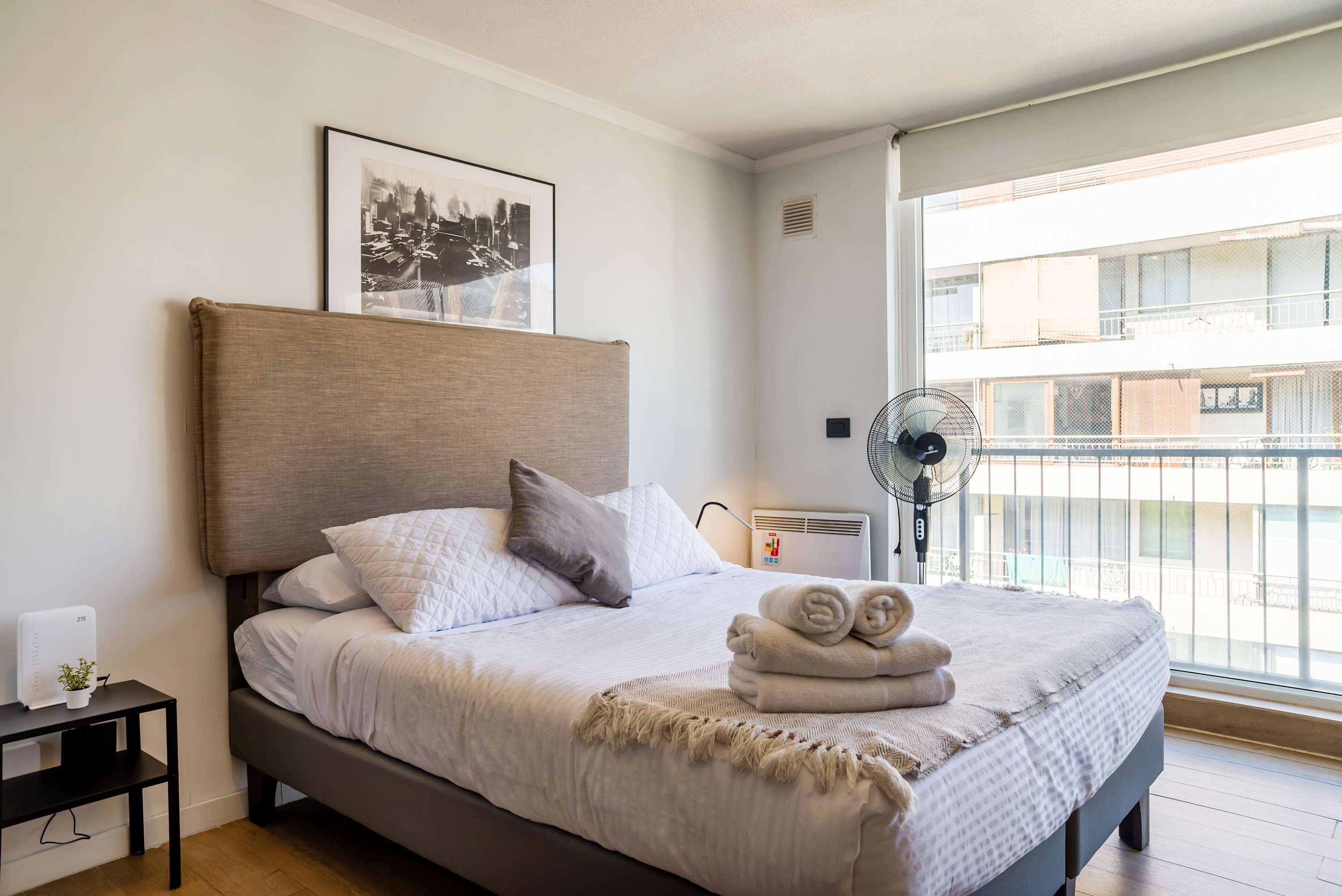 Private studio sleeping area with a neatly made double bed, soft layered linens, rolled towels, and bright windows overlooking the city.