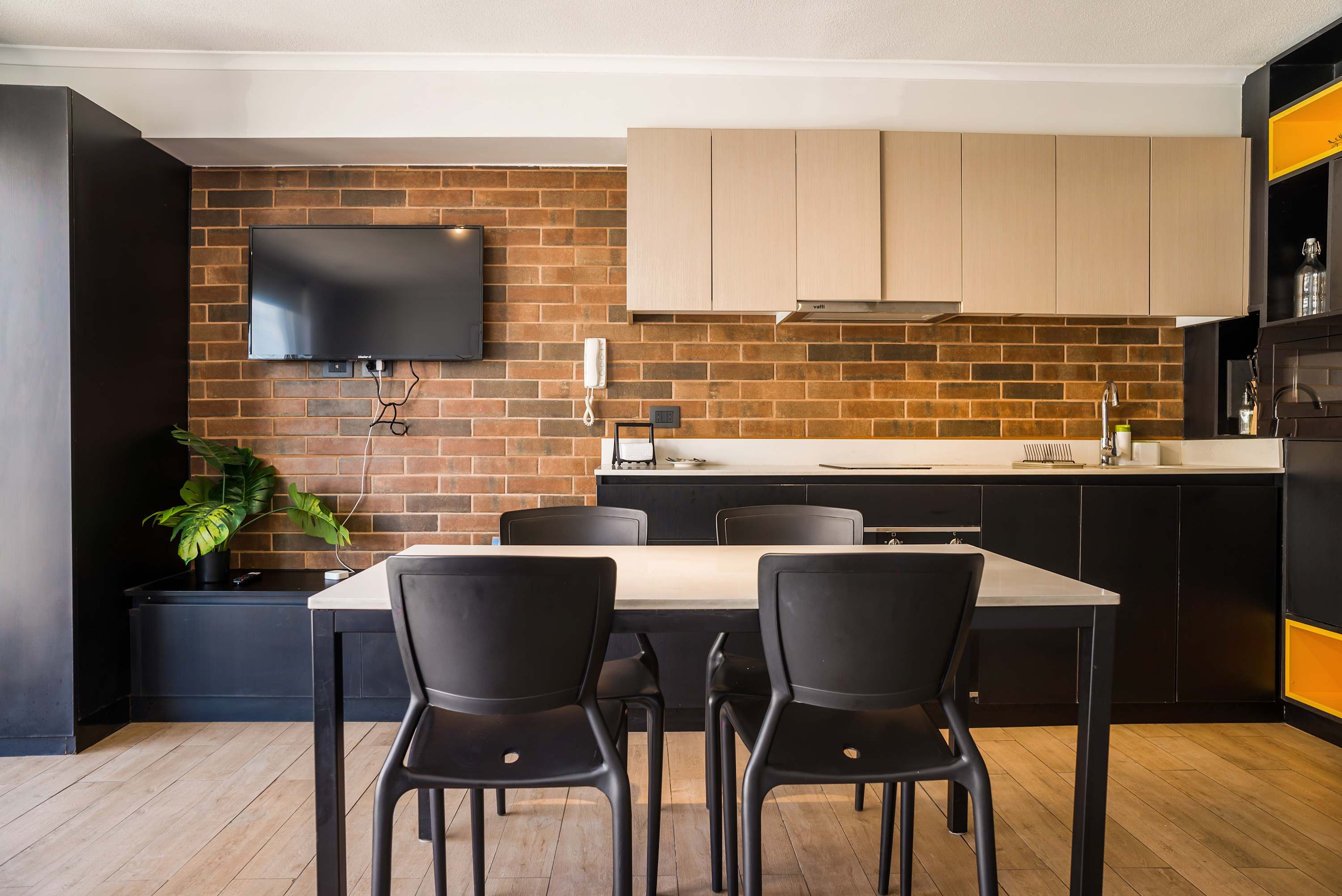 Private kitchen and dining area with a warm brick backsplash, modern black cabinetry, and seating for four beside a wall-mounted TV.