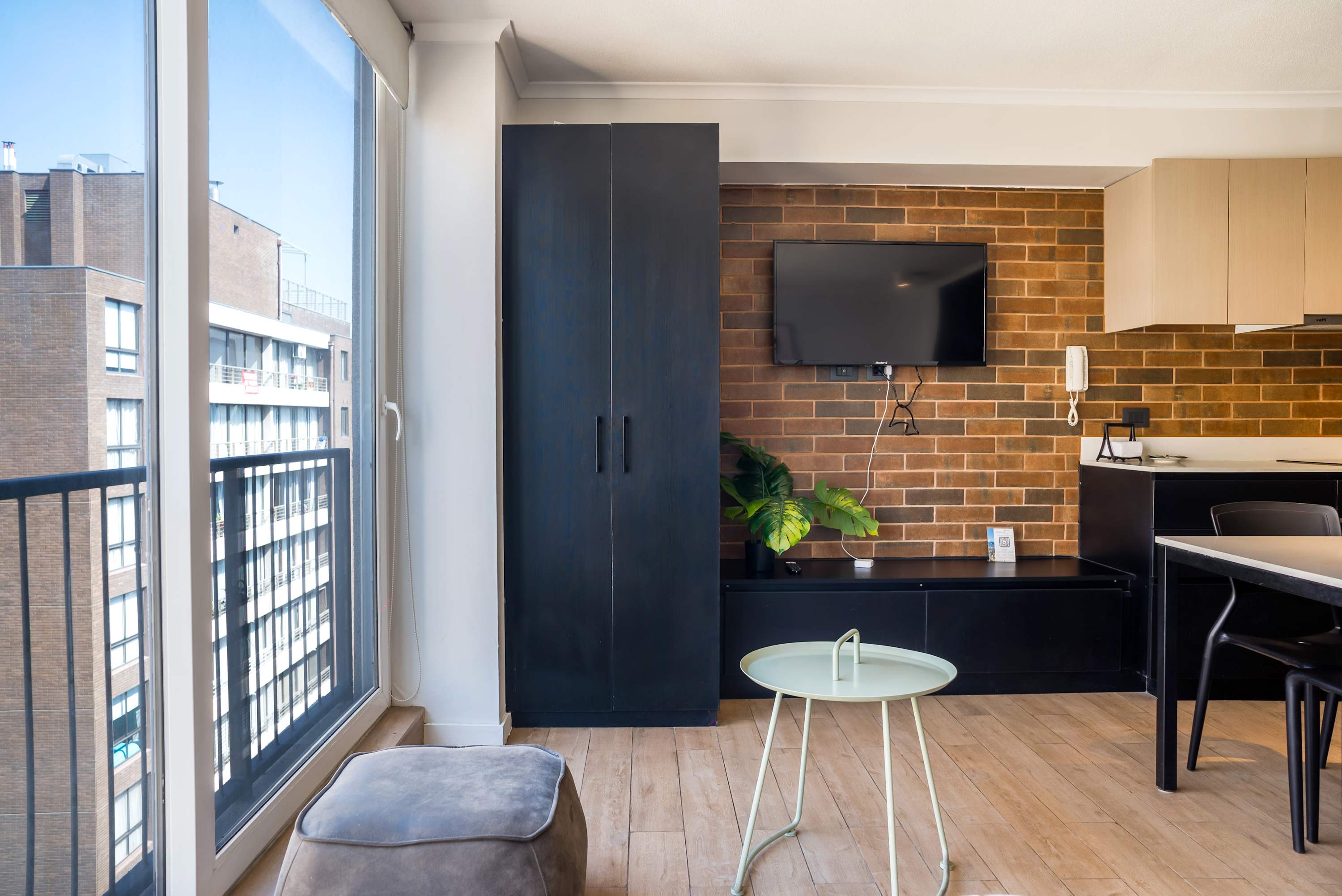 Bright studio living area with floor-to-ceiling windows, a warm brick accent wall, mounted TV, and a compact dining nook.