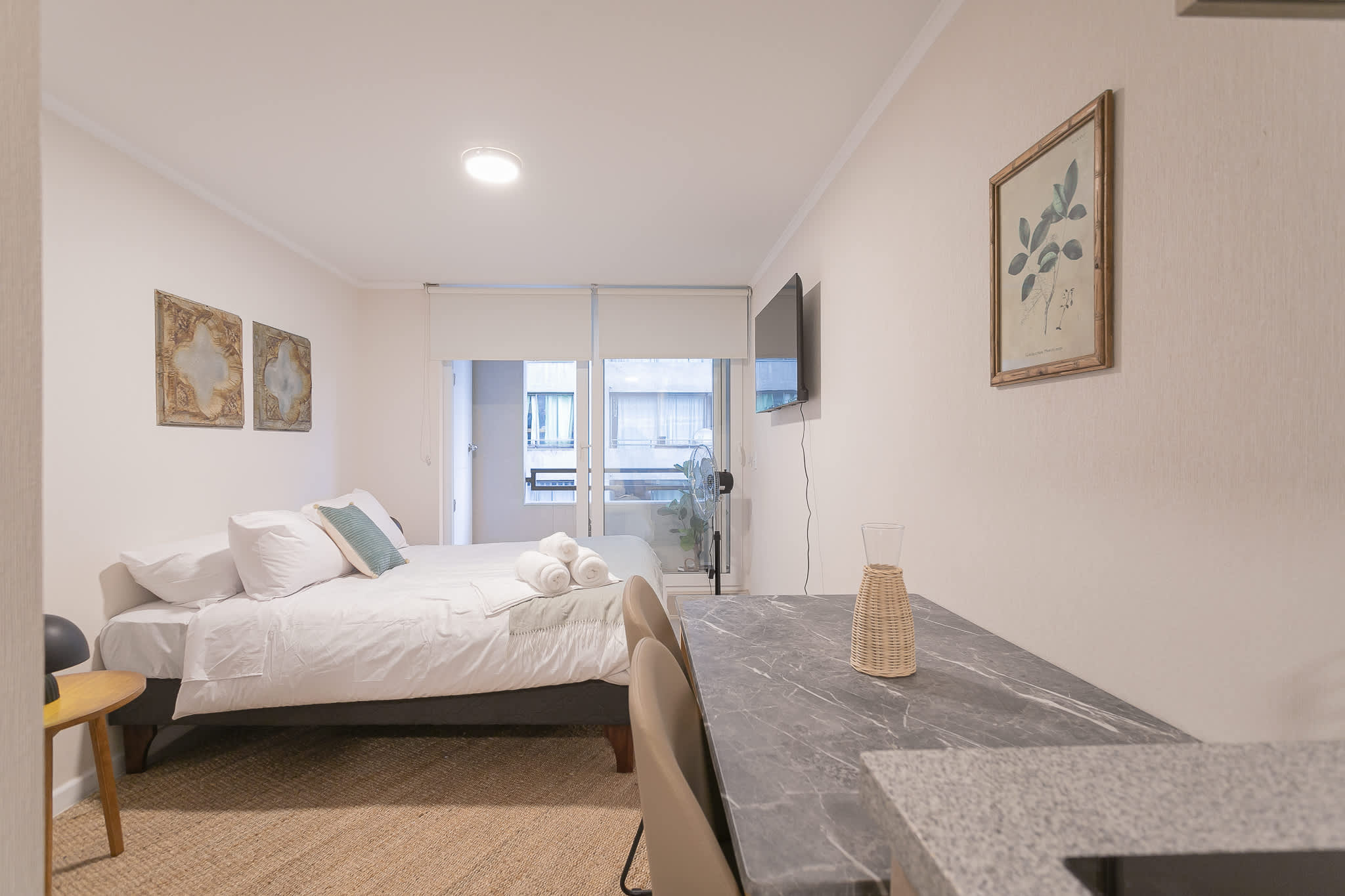 Bright studio bedroom with crisp white bedding, rolled towels, and a marble-look dining table beside the window.