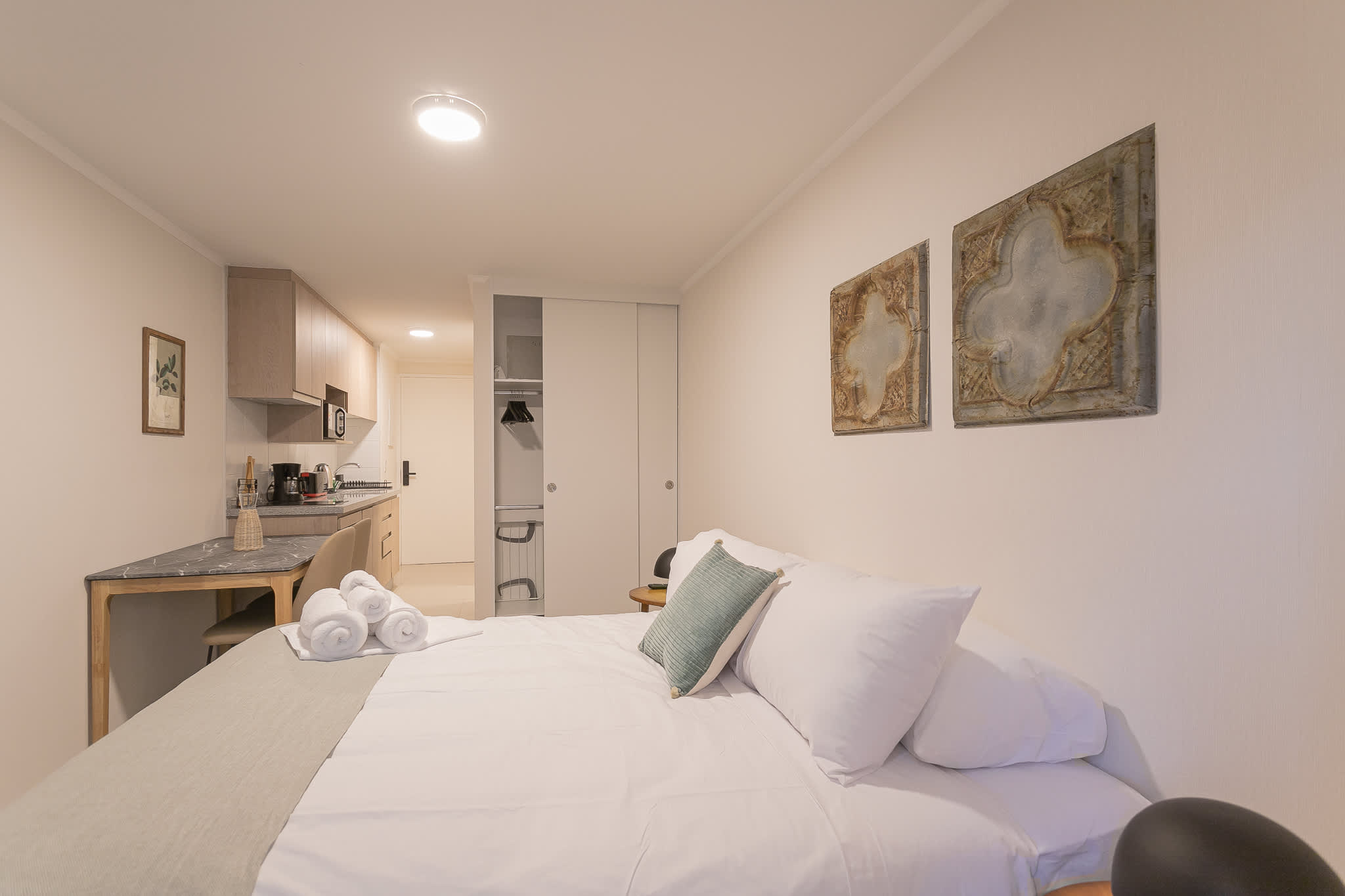 Bright studio sleeping area with crisp white bedding, a soft green accent cushion, and a fully equipped kitchenette just steps away.