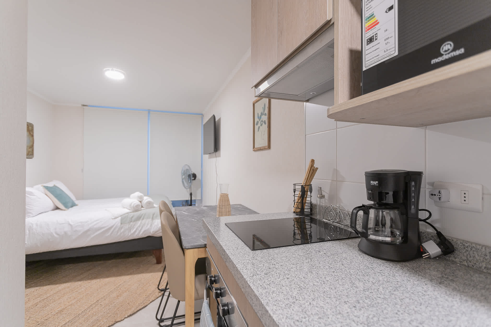 Private kitchen with a black glass cooktop, drip coffee maker, and compact dining counter overlooking the neatly prepared studio sleeping area.