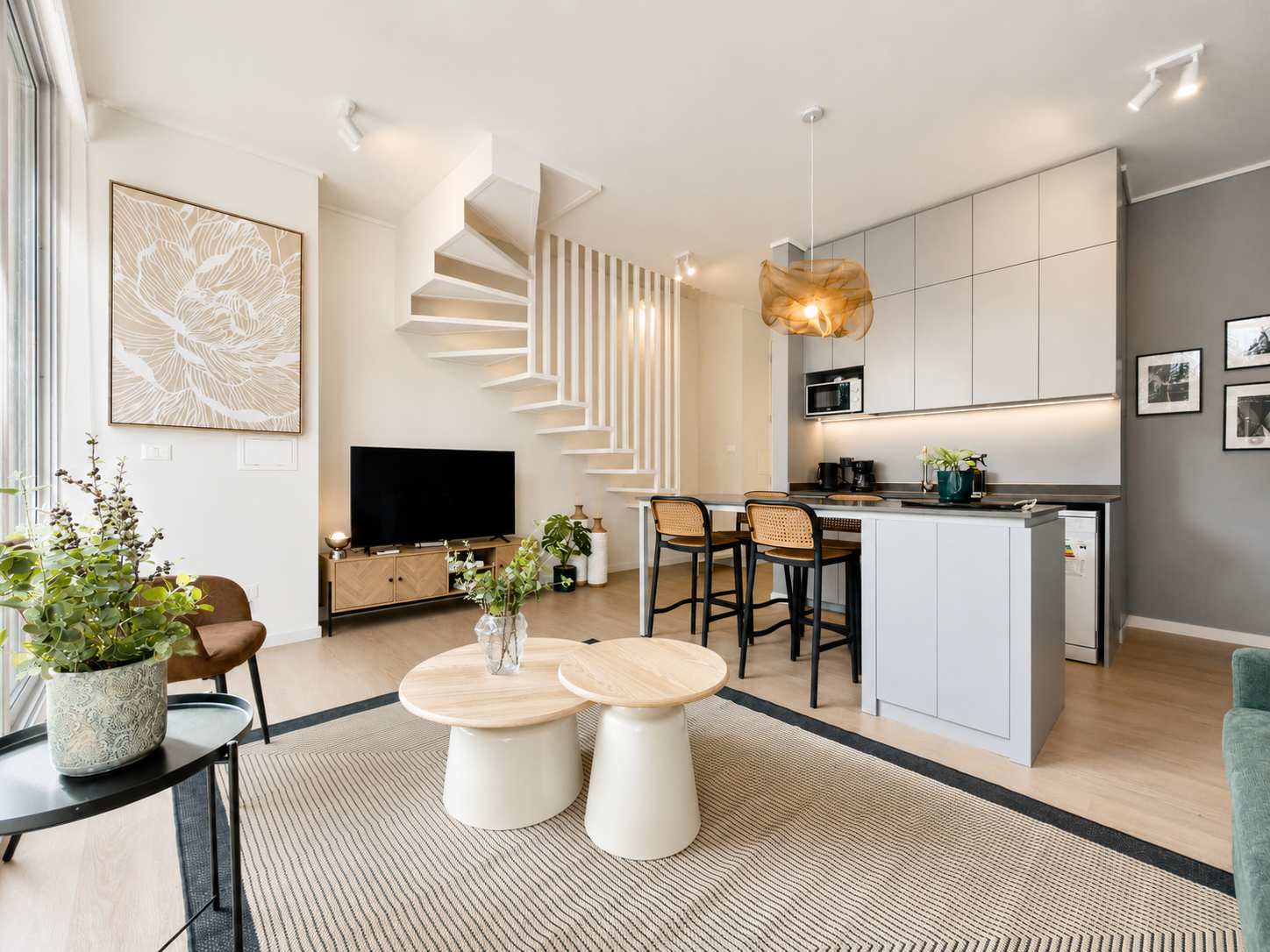 Bright open-plan living room with a sculptural staircase, cozy seating, nesting coffee tables, and a sleek kitchen peninsula with woven bar stools.