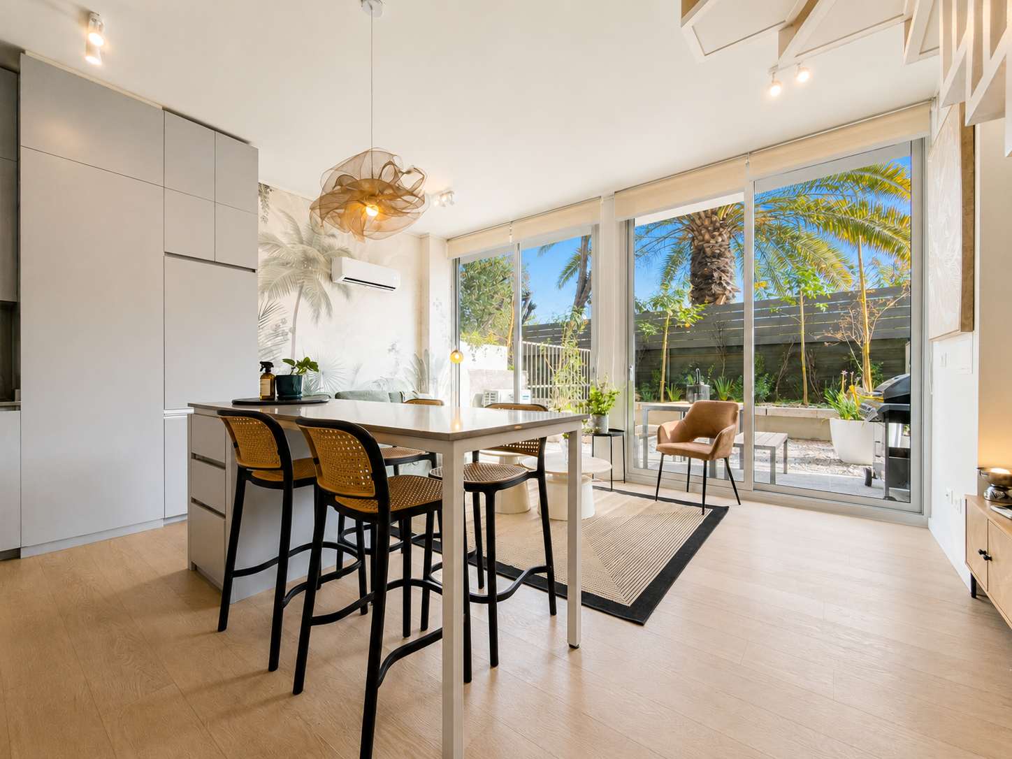 Open-plan kitchen with a sculptural pendant, five-seat island, and glass doors opening to a sunny palm-lined patio.