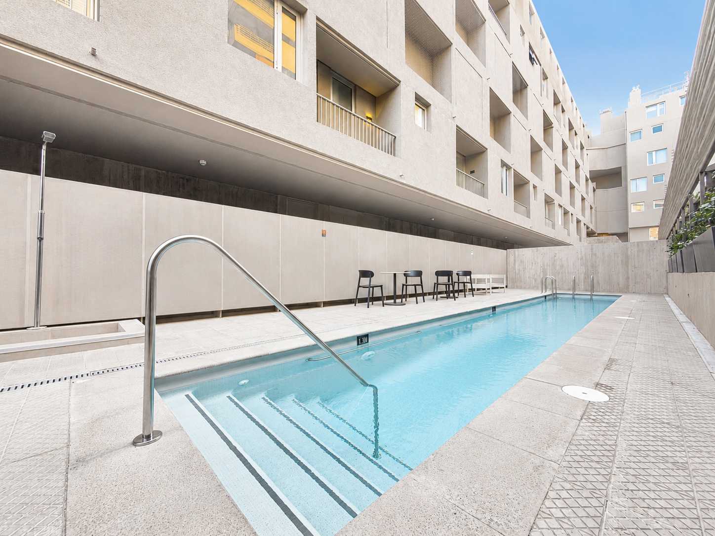 Outdoor swimming pool with clear blue water, stainless-steel entry rails, and courtyard seating surrounded by modern apartment architecture.