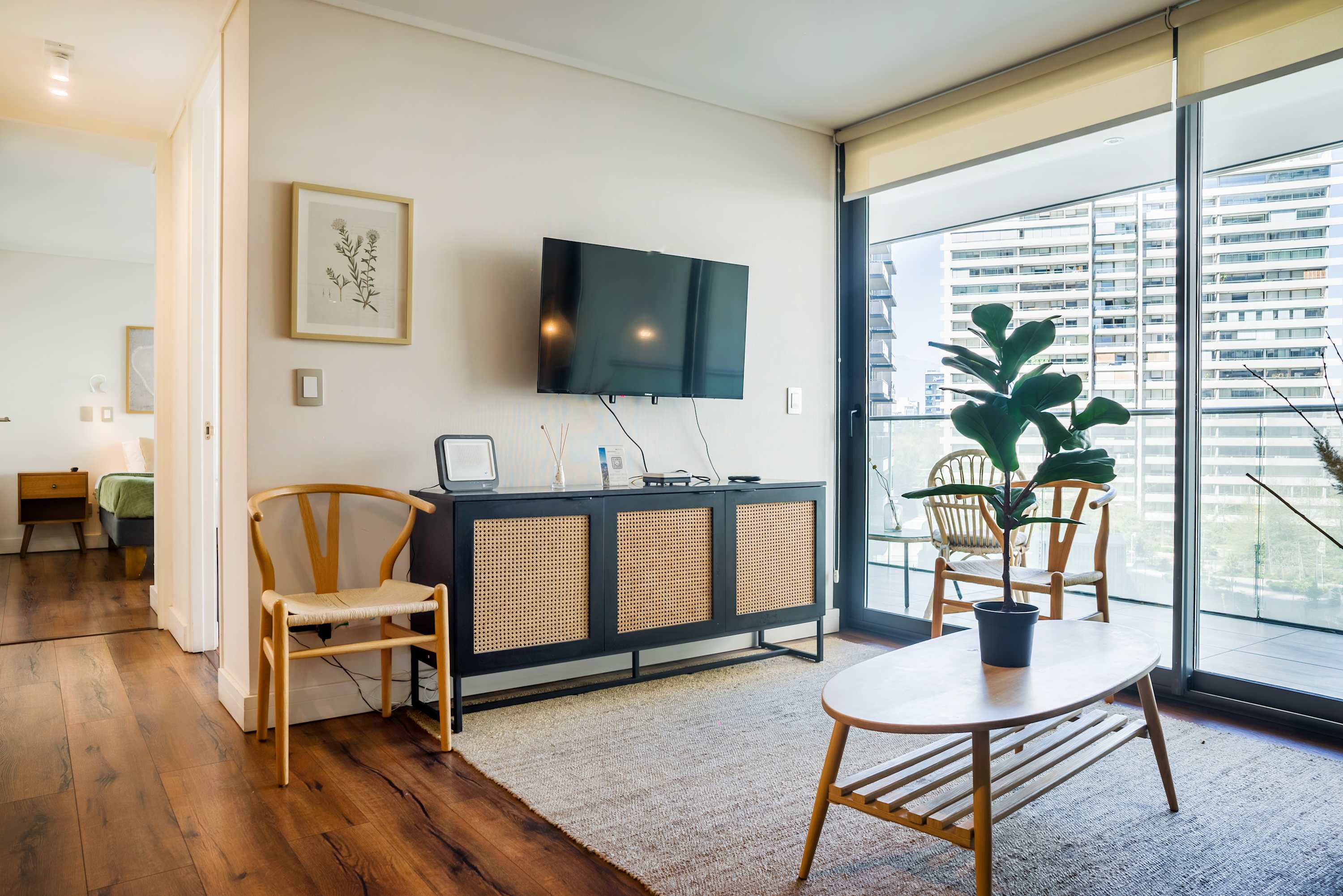 Private living room with a wall-mounted TV, cane-front media console, and floor-to-ceiling windows opening to a bright city-view balcony.
