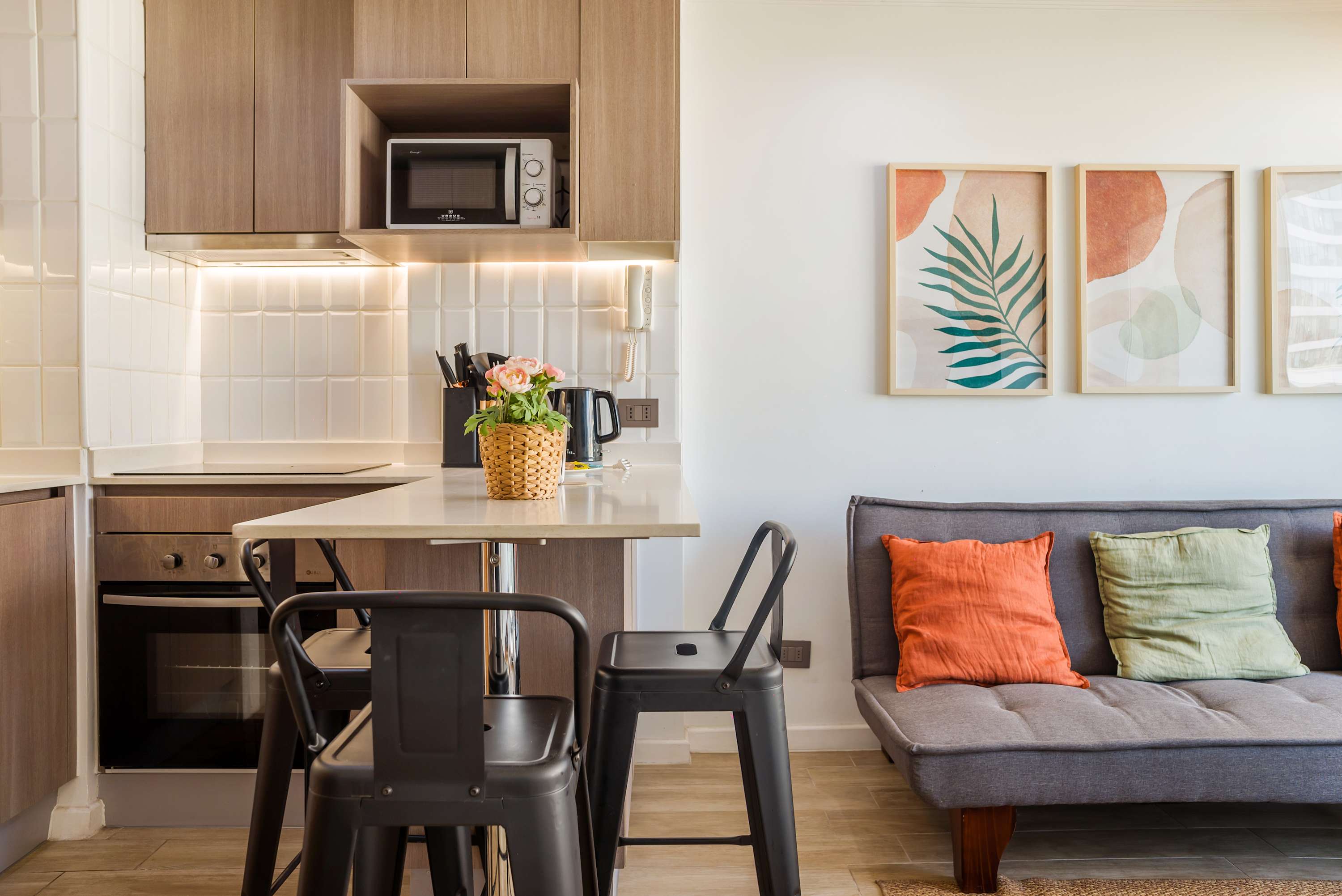 Compact studio living area with a practical kitchenette, bar dining counter, and colorful botanical artwork beside a cozy gray sofa.