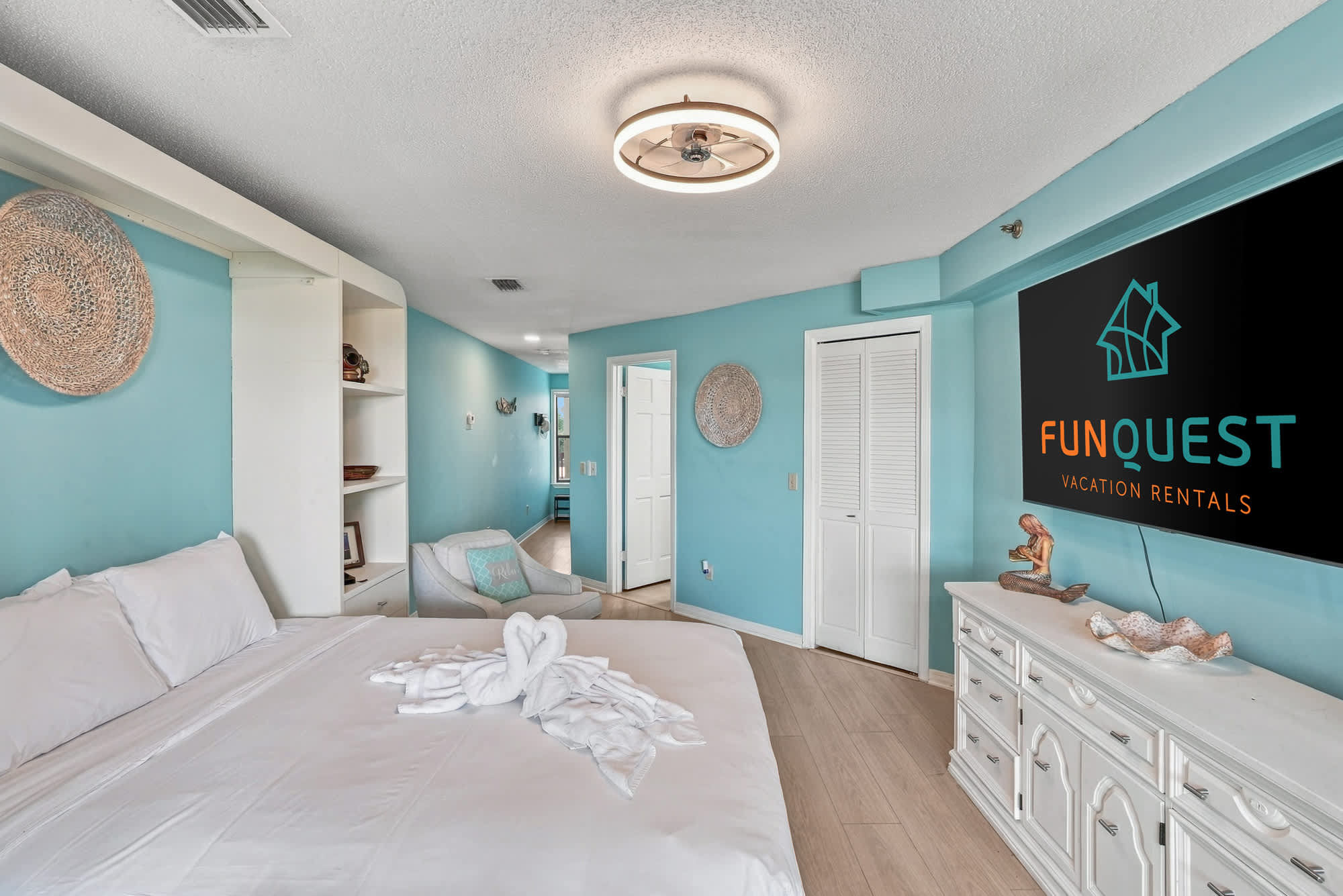 St Thomas SQ 1209C – Bedroom with light blue walls, king bed, decorative towel art, reading chair, and TV on the wall.