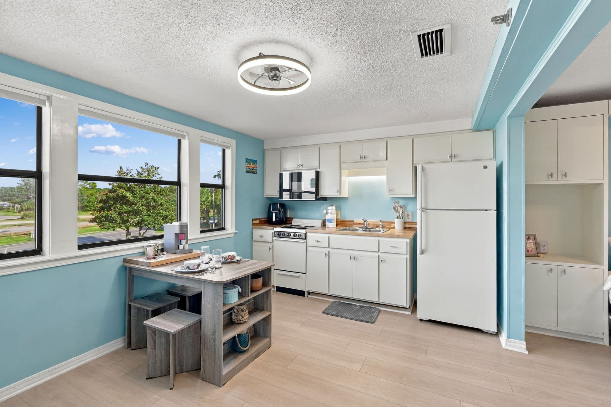 St Thomas SQ 1209C – Bright kitchen with dining area, white appliances, and large windows showing green outdoor view.