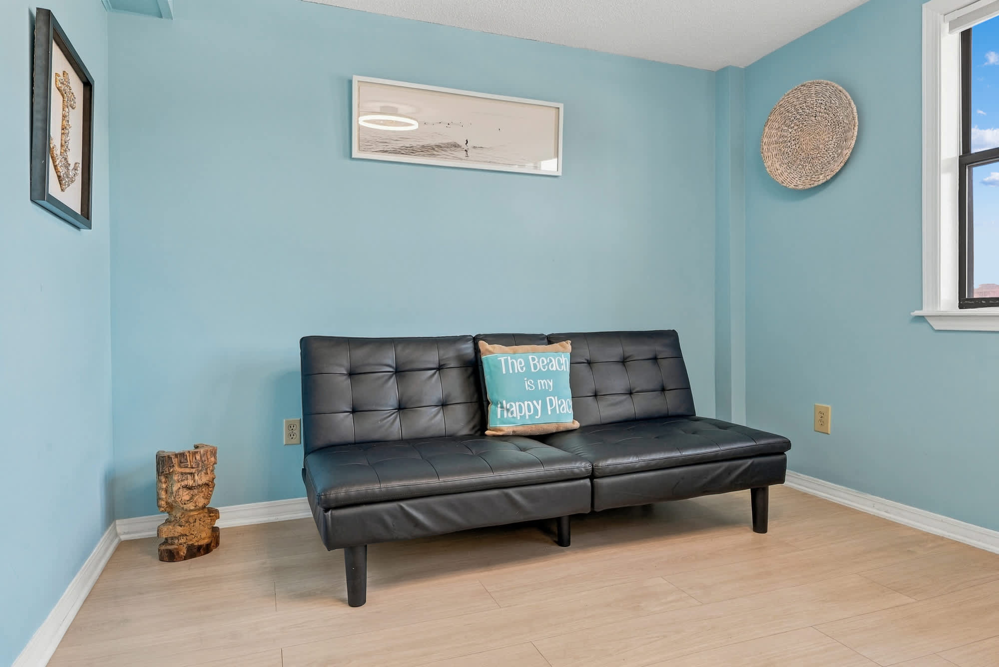 St Thomas SQ 1209C – Living room with light blue walls, black futon, decorative pillow, and wooden accent piece.