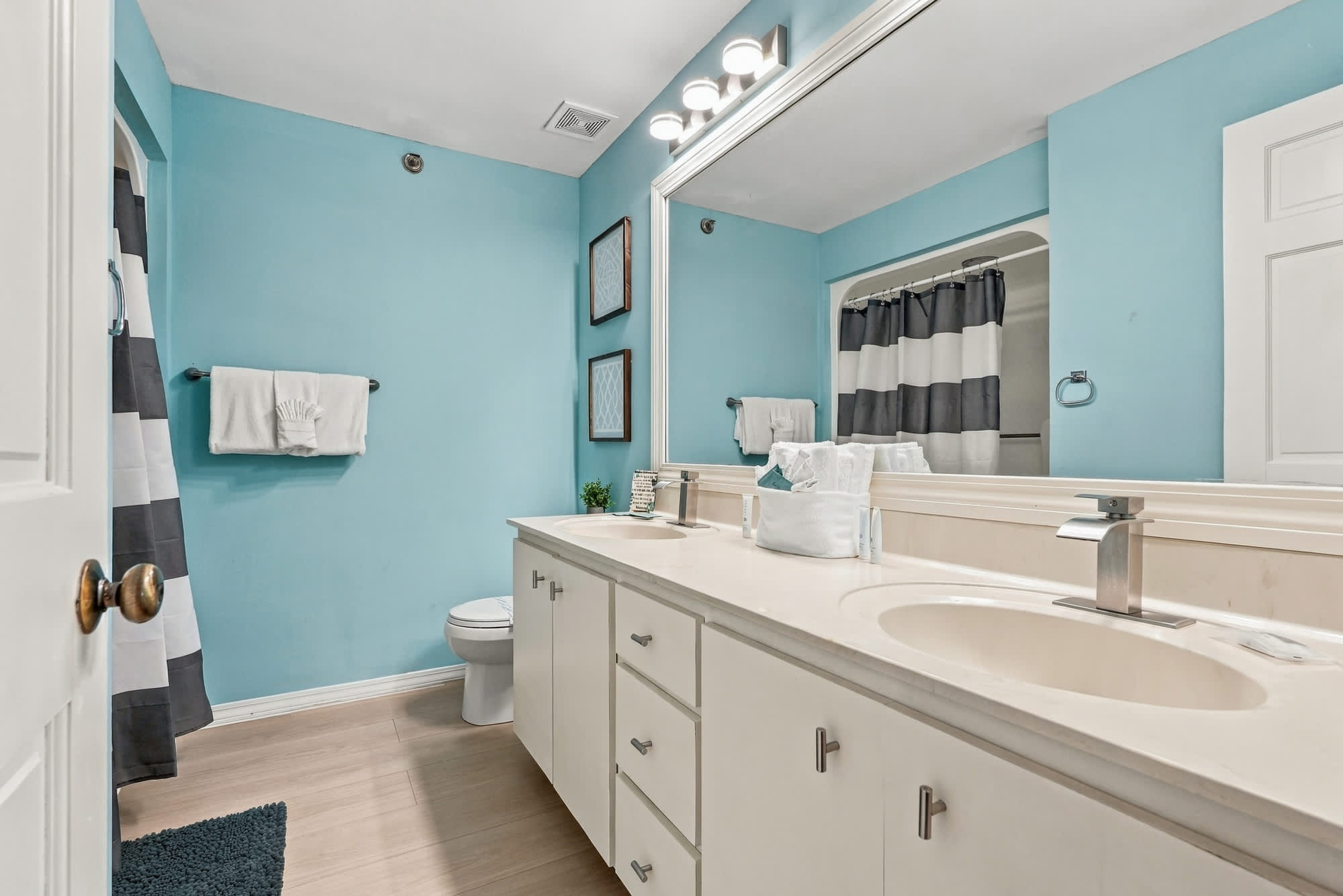 St Thomas SQ 1209C – Bright bathroom with light blue walls, double sinks, and shower curtain in black and white stripes.