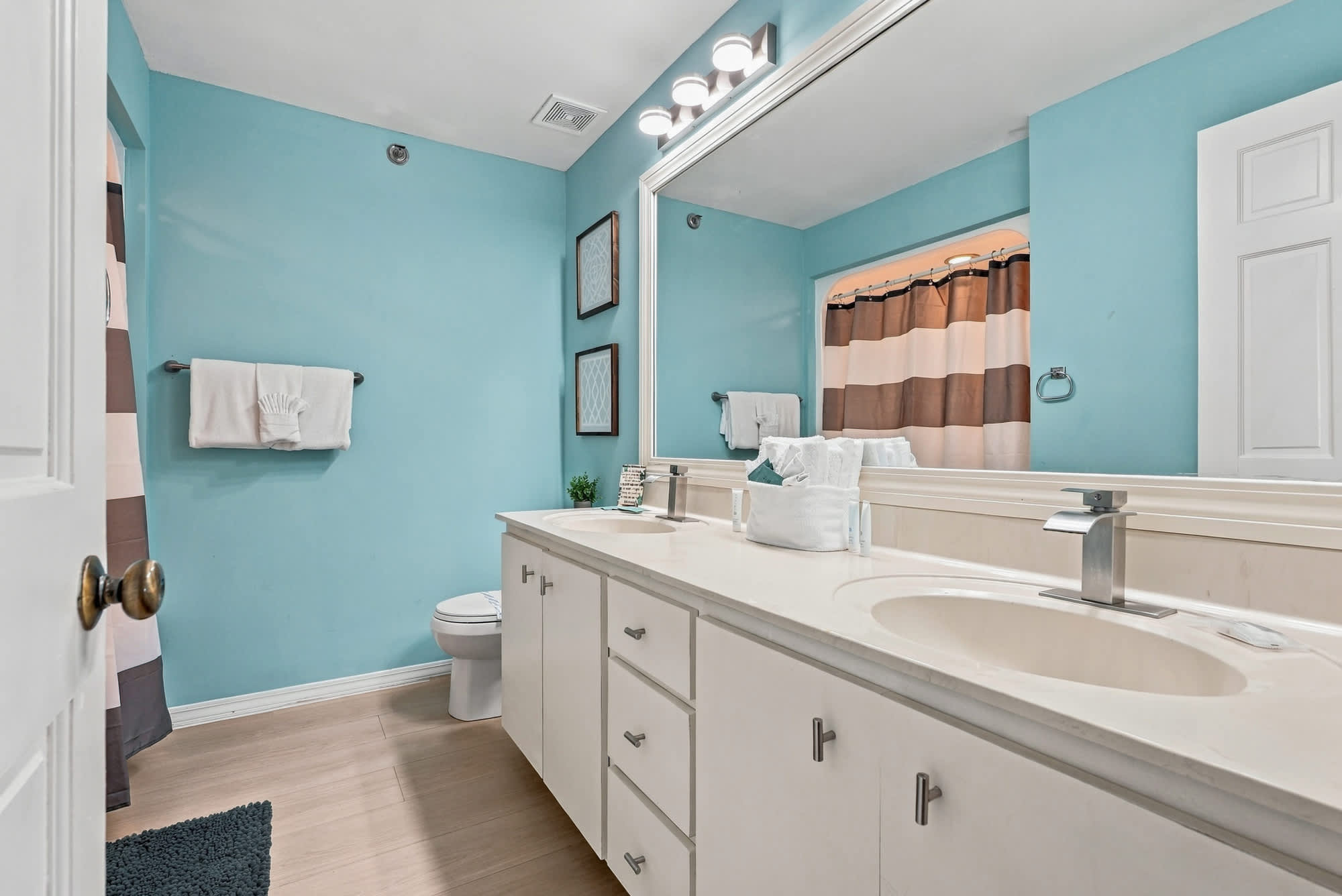 St Thomas SQ 1209C – Bright bathroom with dual sinks, blue walls, and a shower curtain featuring brown and white stripes.