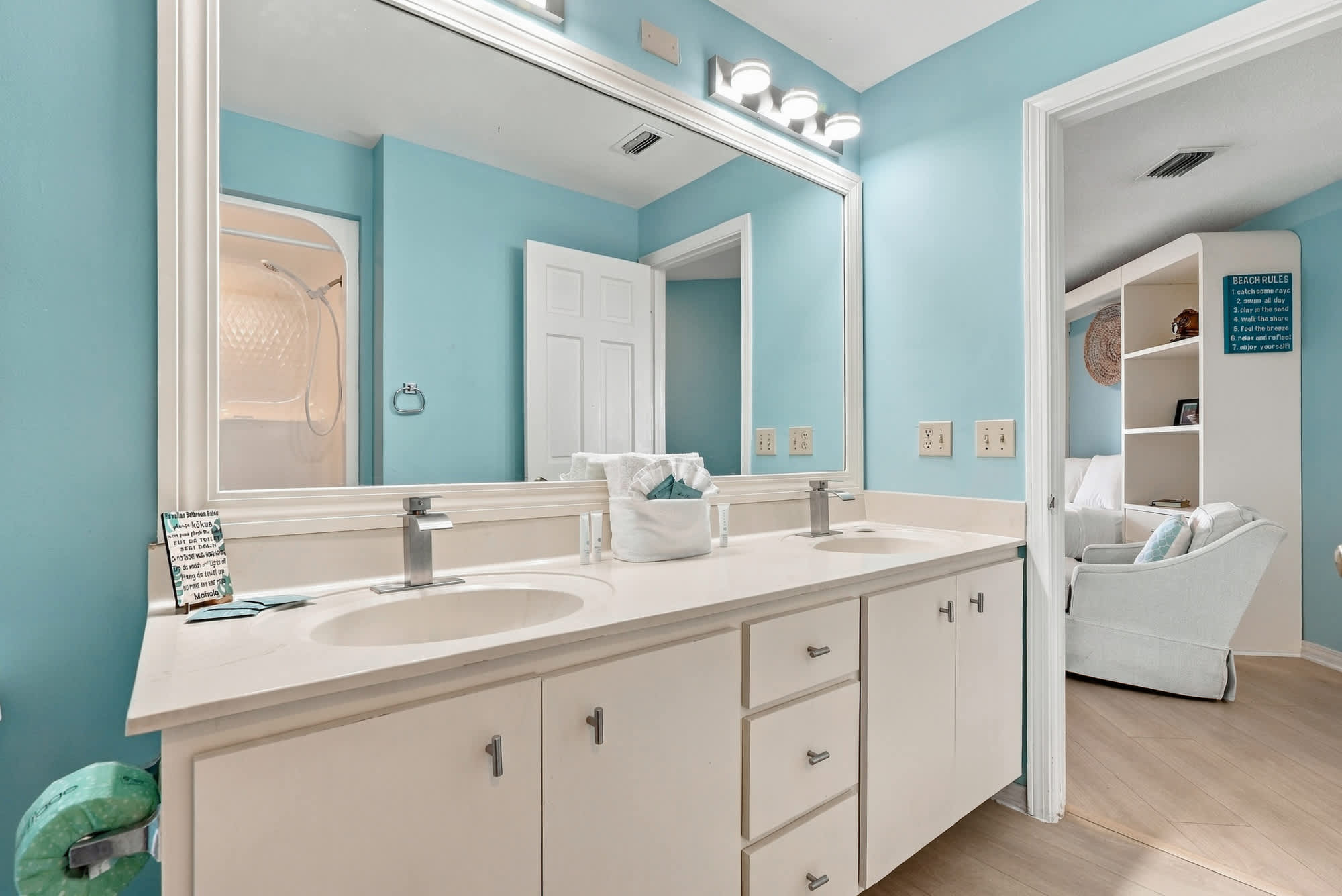 St Thomas SQ 1209C – Bathroom with double sink, light blue walls, and a large mirror, adjoining a living area with a white chair.