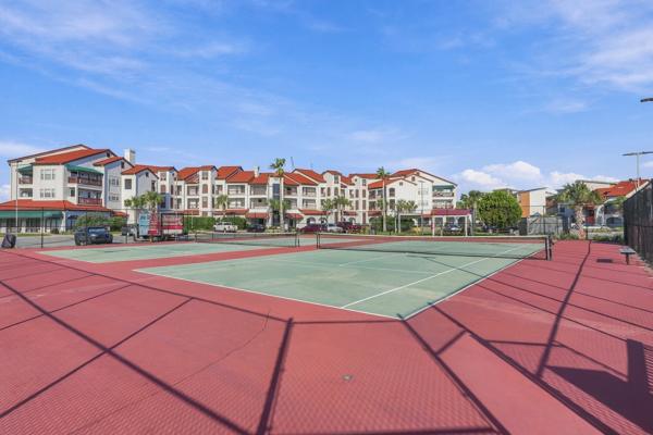 St Thomas SQ 1302B – Tennis court with red surface, surrounded by residential buildings and green landscaping under a clear blue sky.