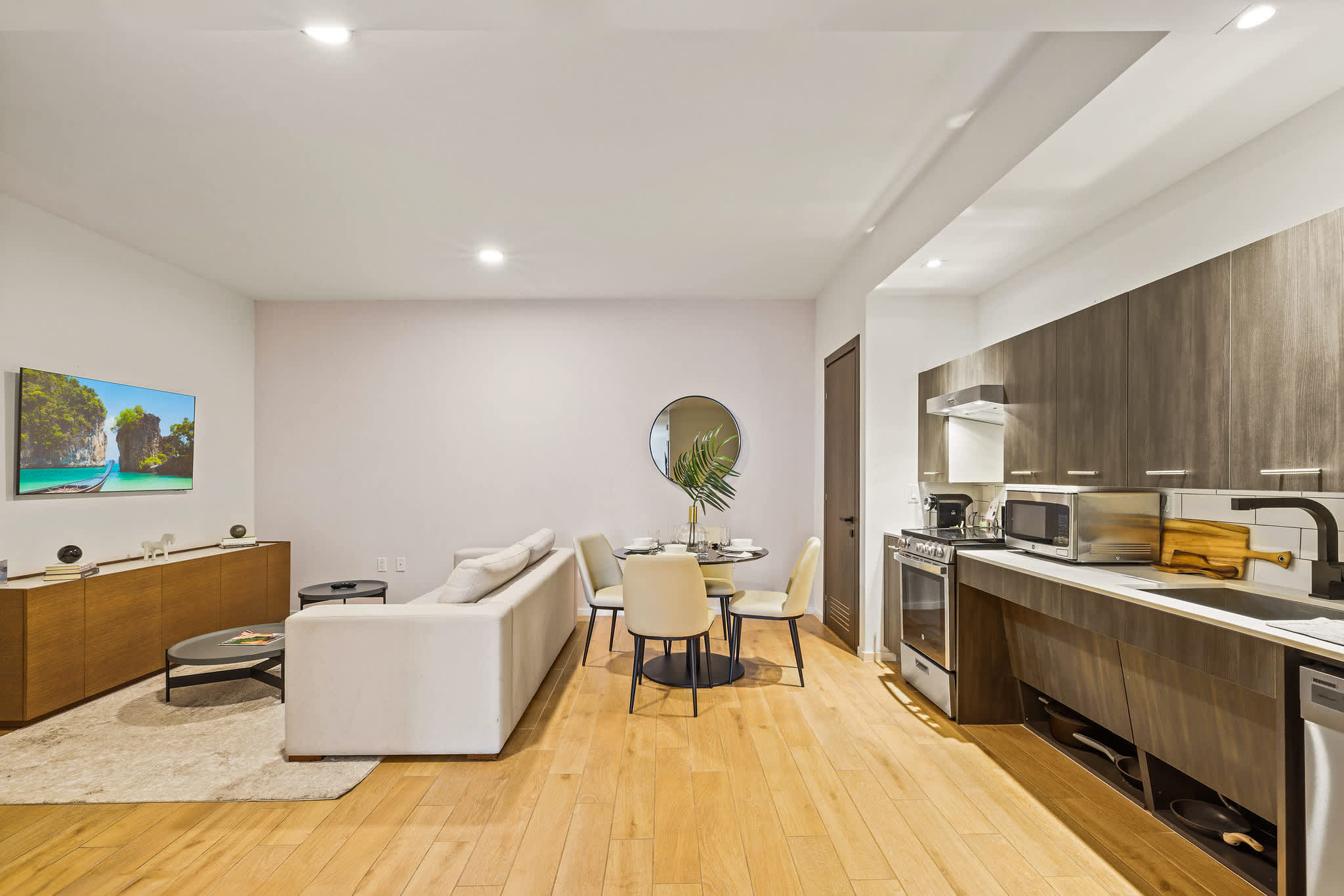 Open-plan living room with a tropical-view TV, dining area for five, and a fully equipped modern kitchen with warm wood cabinetry.