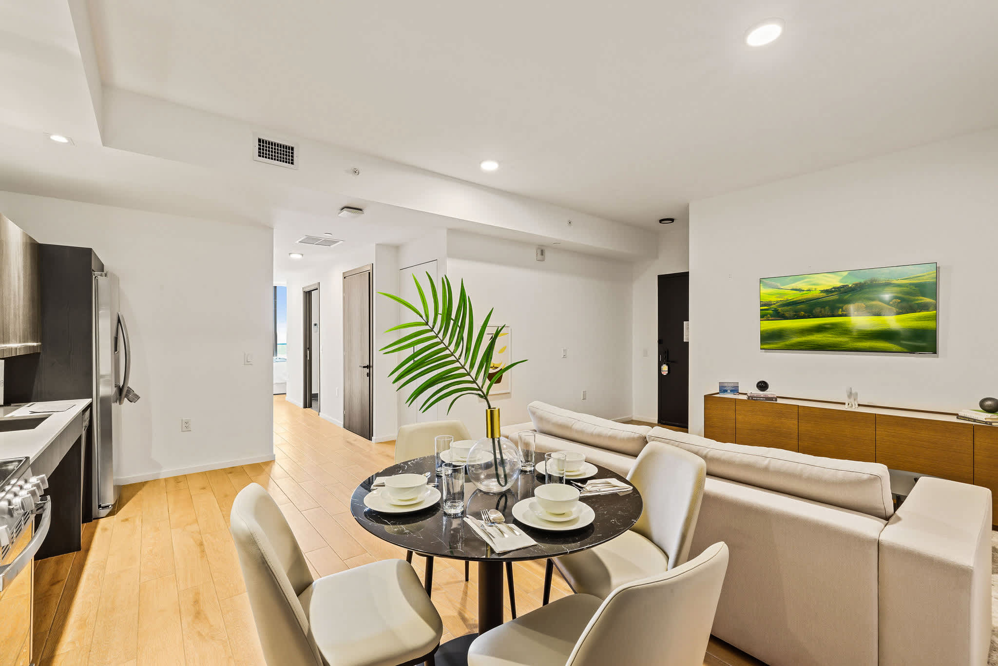 Open-plan living room with a round dining table, cream seating, warm wood floors, and a wall-mounted TV overlooking a landscaped media console.