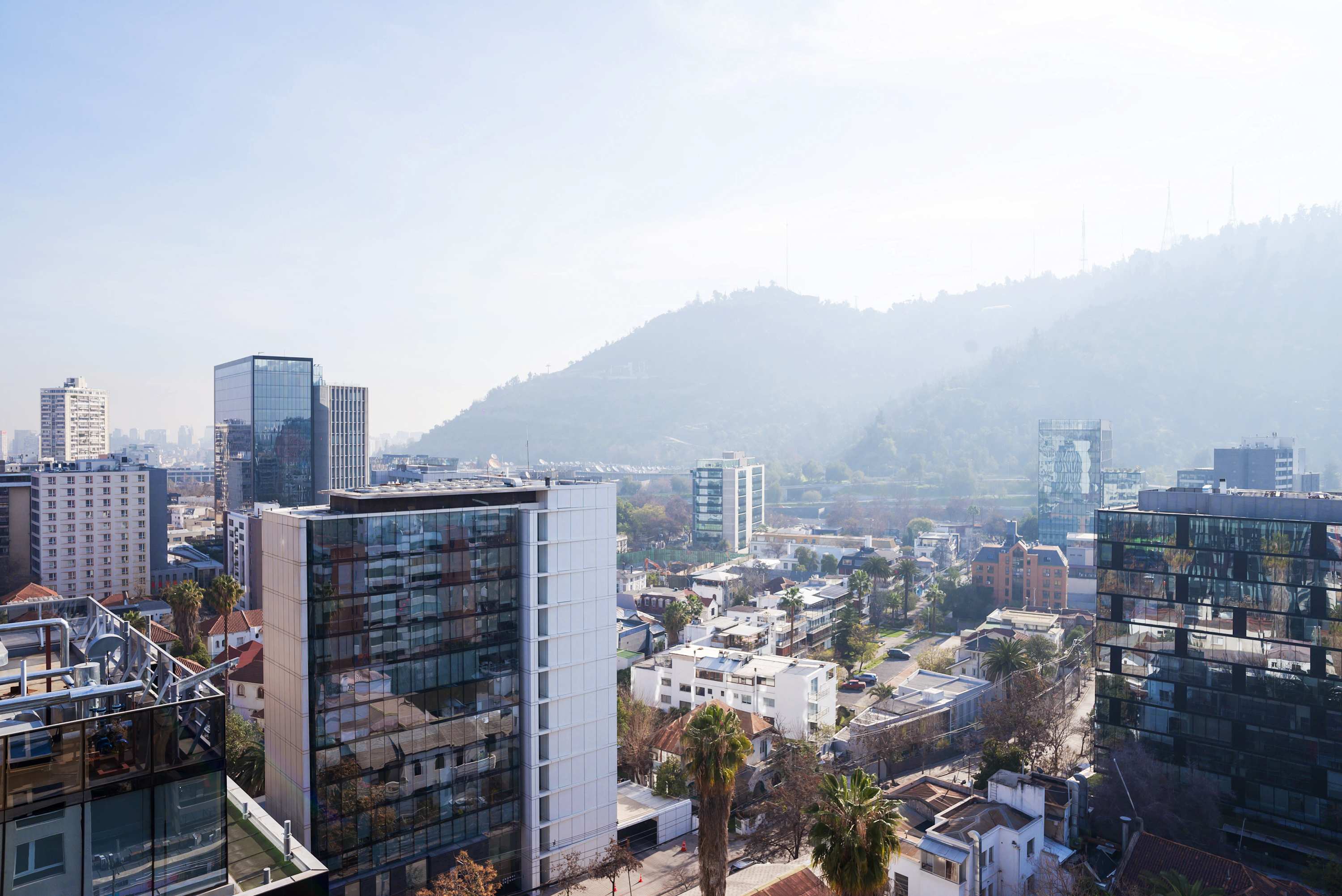 Panoramic Santiago cityscape with glass towers, leafy streets, terracotta rooftops, and a green hillside stretching across the horizon.