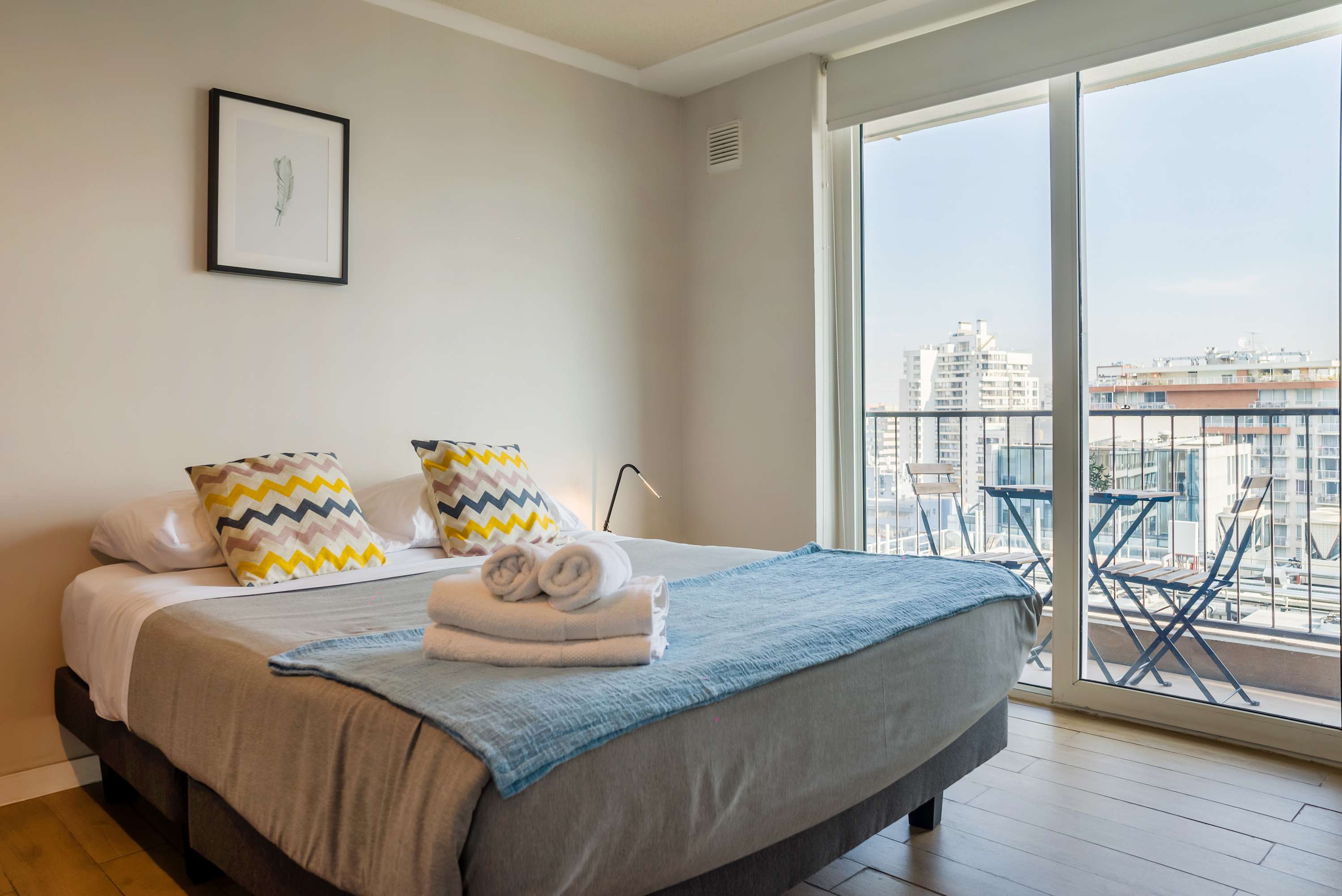 Bright bedroom with a comfortable double bed, colorful patterned pillows, and a private balcony overlooking the city.