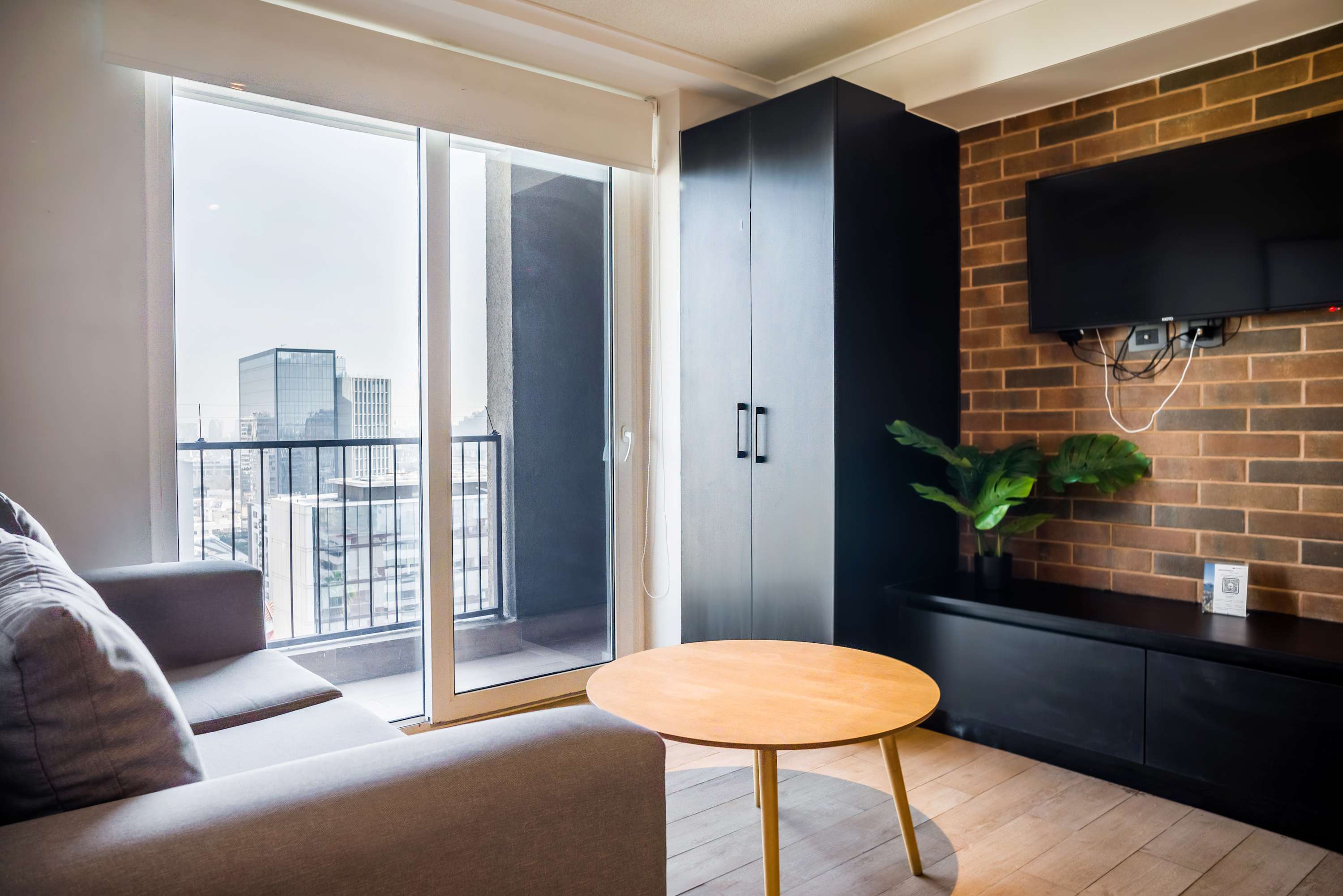 Contemporary living room with a cozy gray sofa, round wood coffee table, and city views through the floor-to-ceiling balcony windows.