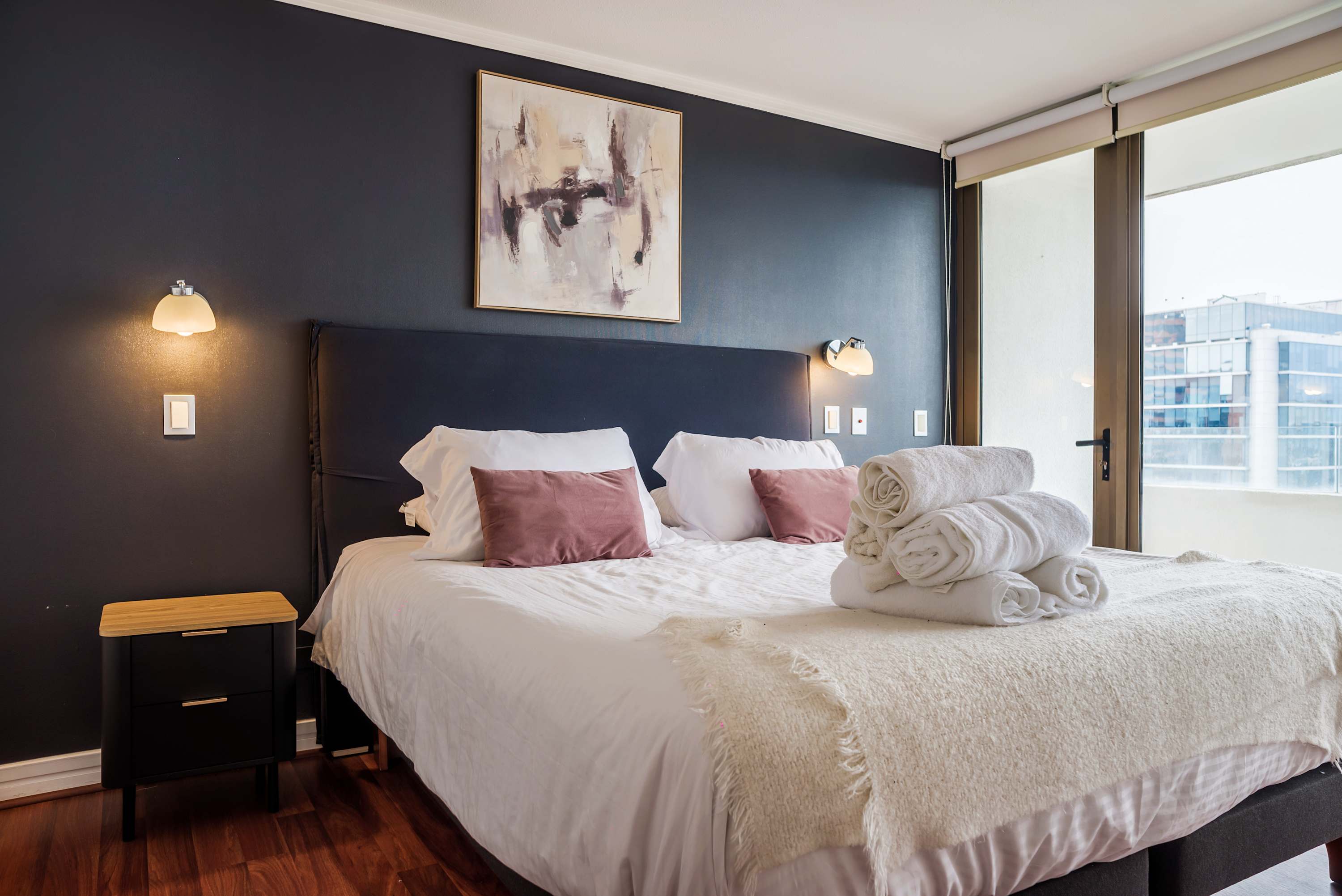 Comfortable bedroom with crisp white bedding, blush-pink cushions, warm reading lights, and a bright urban view through the large window.