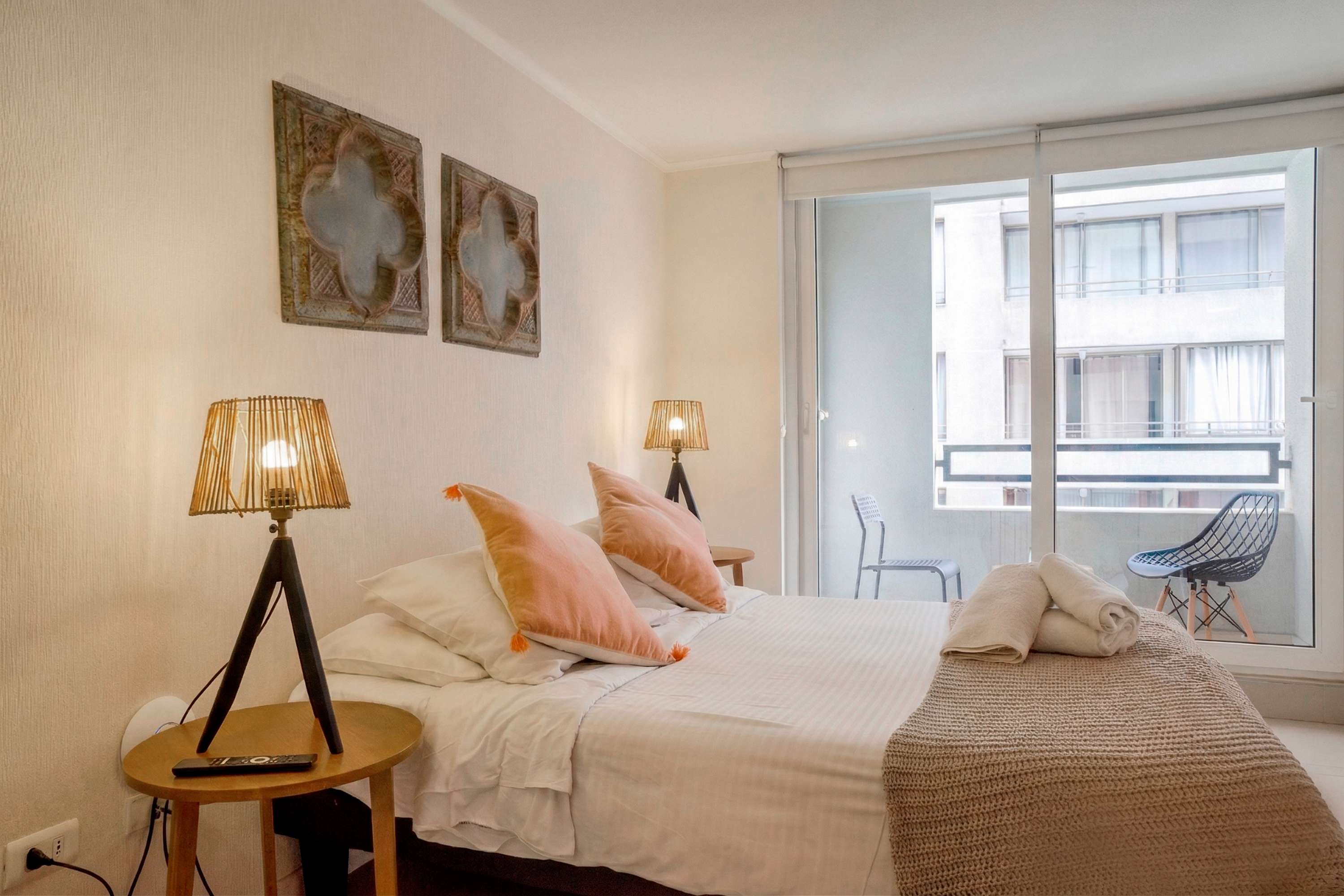 Private bedroom with blush accents, woven lamps, and a bright balcony overlooking the surrounding city buildings.