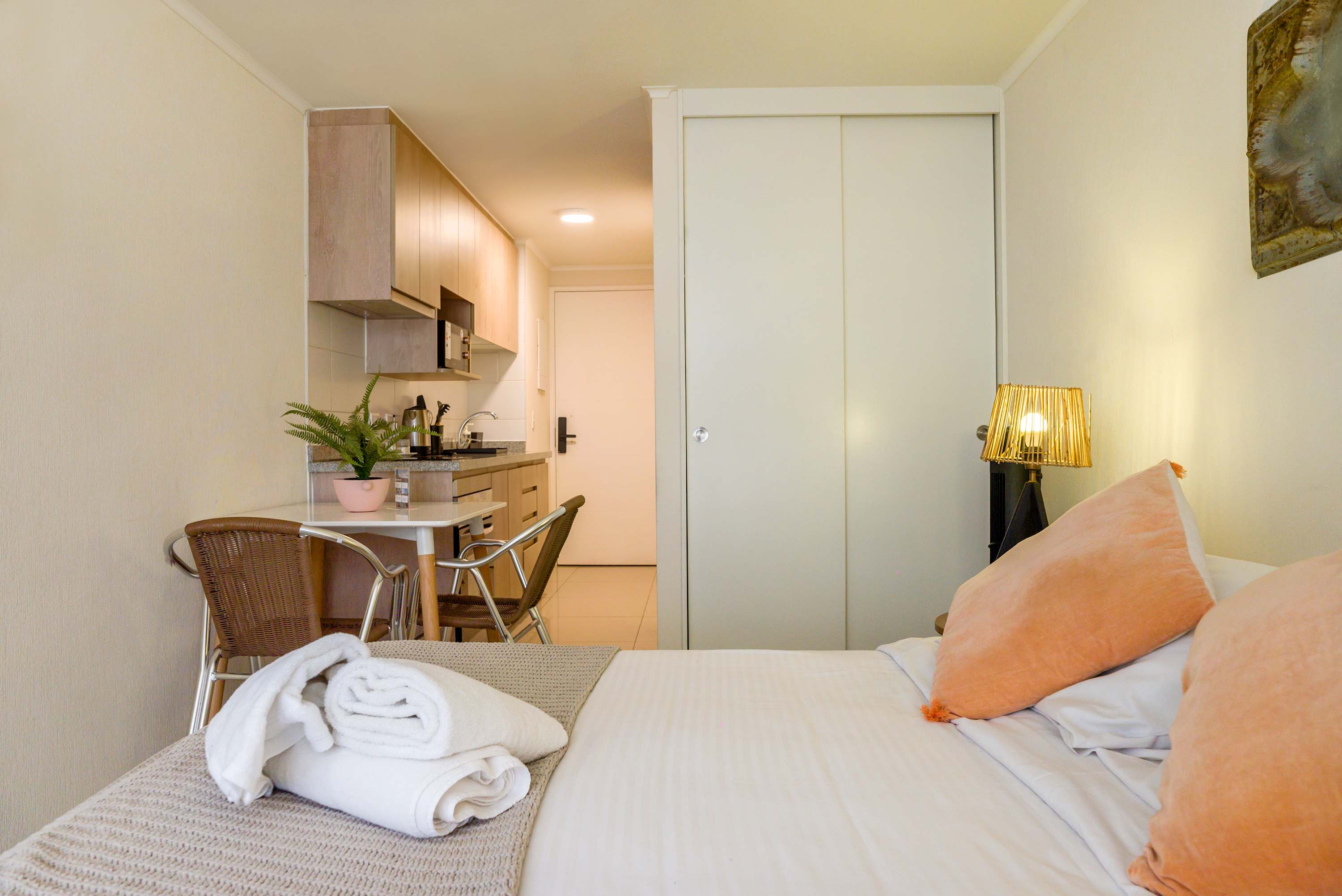 Studio bedroom with soft peach bedding, a compact equipped kitchenette, and a two-person dining nook.
