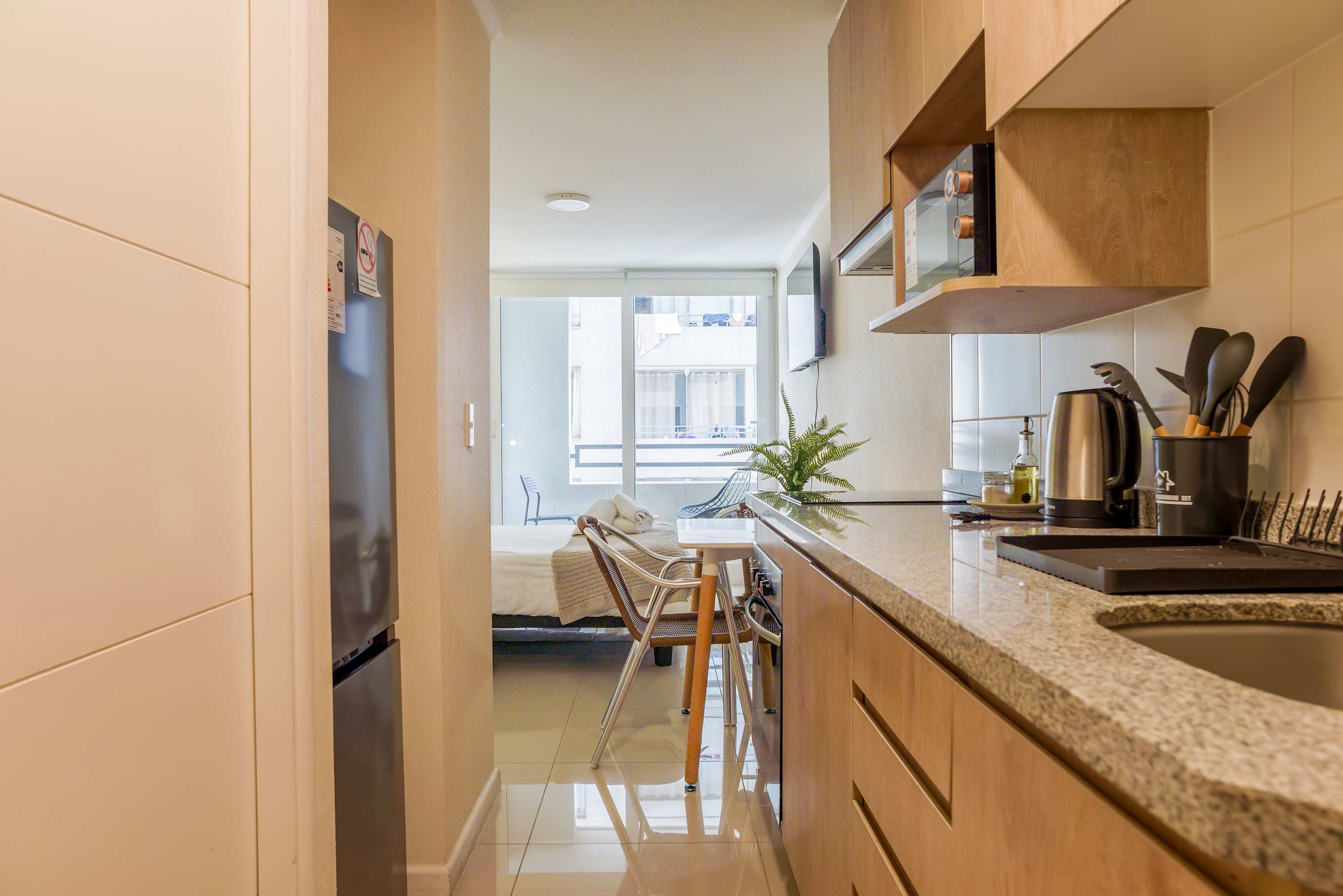 Modern studio kitchen with warm wood cabinetry, a stone countertop, and a breakfast counter opening toward the bright balcony and living area.